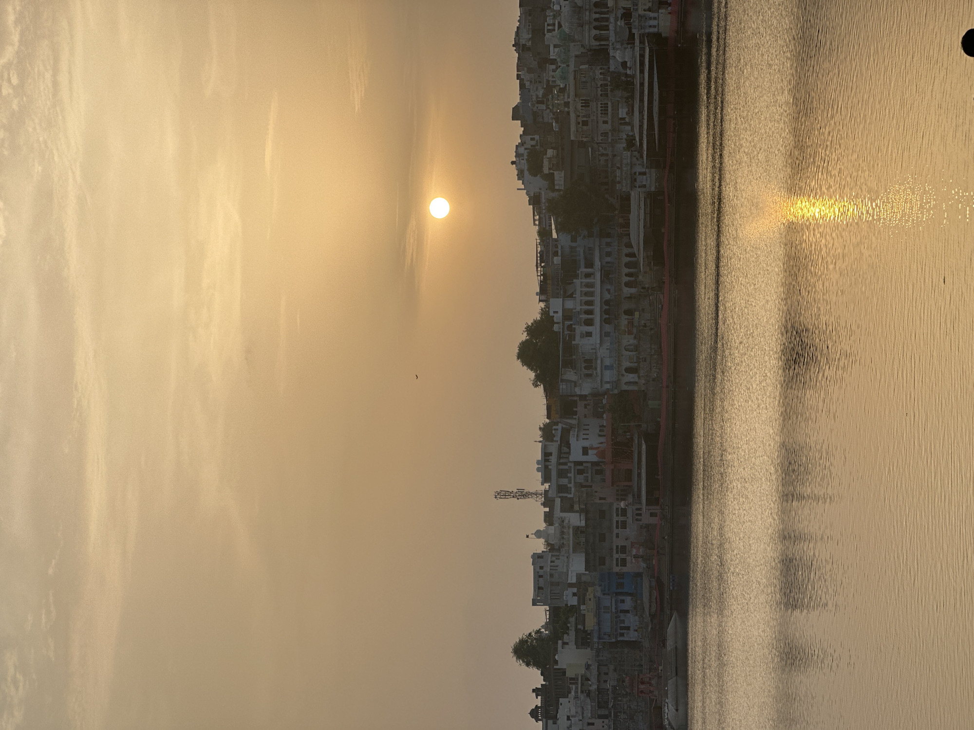 The image captures a serene sunset over Pushkar Lake in Pushkar, India. The sky is a soft gradient of orange and yellow, with faint clouds scattered across it. The sun, a bright, low orb, casts a golden glow on the scene.
The foreground is dominated by the calm waters of Pushkar Lake, reflecting the warm hues of the sky and the cityscape. Ripples on the water's surface create a shimmering effect, with a prominent sun reflection extending from the sun's position towards the viewer.
In the mid-ground, a collection of buildings lines the opposite bank of the lake. These structures, varying in height and architectural style, display a mix of white, beige, and pastel colors, with some displaying more vibrant hues of blue and red. Architectural features like arched windows and balconies are visible. A cellular tower is prominently positioned among the buildings. The lakeside appears to be a developed area with structures close to the water's edge.
A faint silhouette of a bird can be seen in the sky above the buildings, adding a touch of life to the expansive sky. The overall impression is one of tranquility and the beauty of a sunset over a sacred Indian city.