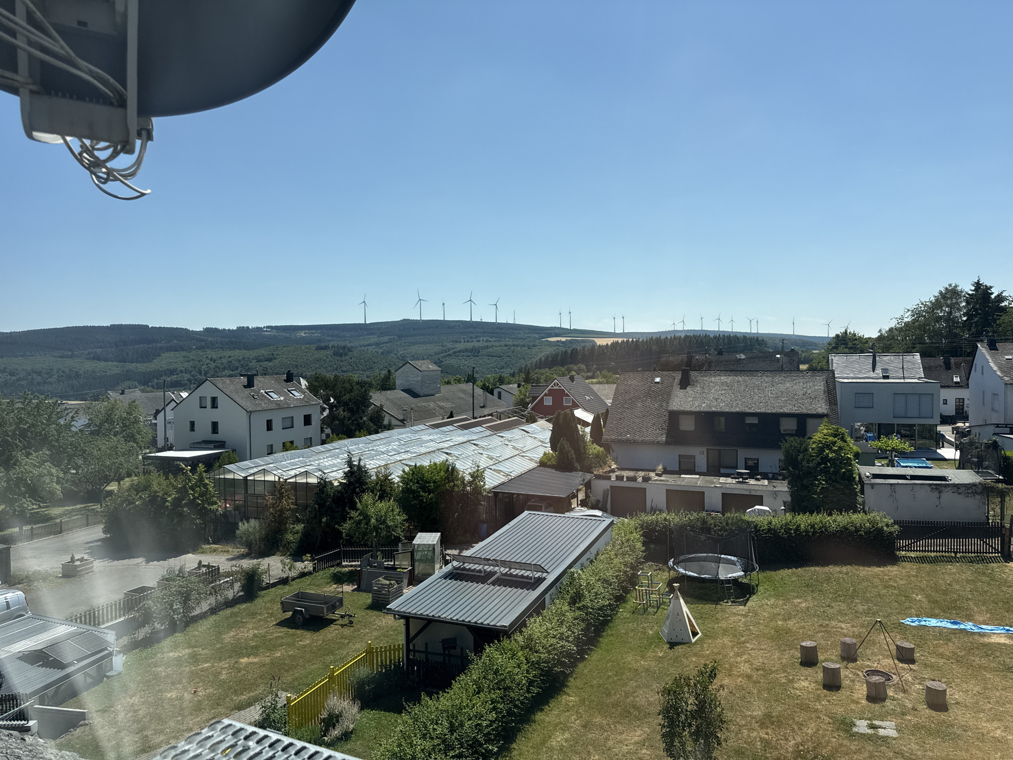 The image shows a view of a residential area in Monzelfeld, Germany, on a clear, sunny day. In the foreground, a large satellite dish is visible, partially obscuring the sky. The middle ground features a collection of houses with dark roofs, some with solar panels. A greenhouse with a transparent roof is also present. Beyond the houses, a grassy yard contains a trampoline, a play tent, a fire pit with log seating, and a small blue tarp. In the background, a series of rolling hills covered in dense green forest stretch to the horizon. Several wind turbines are clearly visible on the crests of these hills. The sky is a vibrant, cloudless blue.