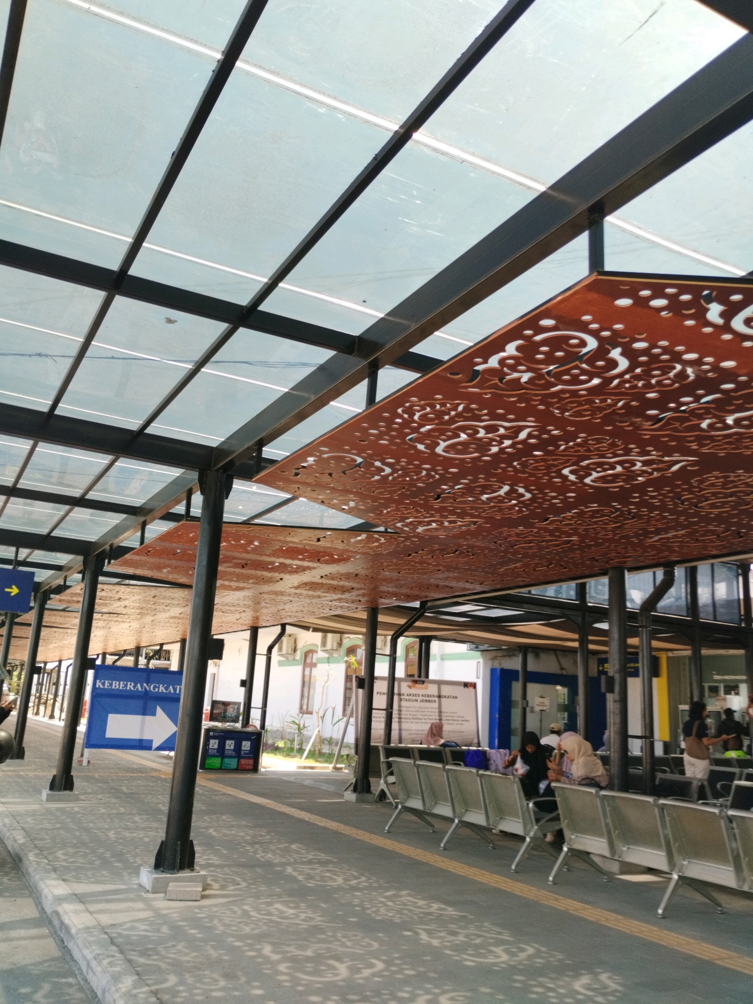 This is an outdoor scene at the Jember train station in Indonesia. The image captures a waiting area with a transparent roof supported by black metal beams and pillars. Decorative, perforated panels with intricate patterns are suspended from the roof structure. Rows of silver metal chairs are arranged on a patterned concrete floor. A blue sign with a white arrow and the text "KEBERANGKATAN" (meaning departure) is visible. In the background, there are buildings, some with visible windows and doors, and several people are seated in the waiting area, some looking at their phones, and others appearing to converse. A trash can is also present. The lighting suggests it is daytime, possibly with some overcast conditions due to the diffusion of light through the roof. The overall impression is of a functional and somewhat modern public transportation waiting area.