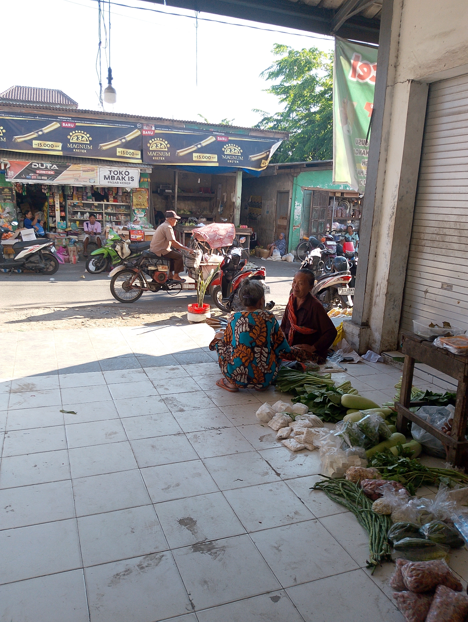 This is a street-level, medium shot image of a market stall in Kendal Rejo, Indonesia. The scene is bustling with activity. In the foreground, two elderly women are seated on the tiled ground, one in a vibrant, patterned dress and the other in a dark brown garment with a red sash. They appear to be interacting, with the woman in brown looking towards the woman in the patterned dress. Piles of fresh vegetables, including long beans, leafy greens, and gourds, are laid out in front of them. Bags of other produce and possibly spices are also visible.
Behind the women, a man on a motorcycle is being pushed or is preparing to depart. The motorcycle has a basket on the front and a canopy for protection from the sun. Further back, several other motorcycles are parked. The market appears to be set up in front of a row of shops. Prominent signage for "DUTA KRETEK" and "MAGNUM KRETEK" are visible, indicating the sale of cigarettes. The shops display a variety of goods on shelves. The environment suggests a typical Indonesian street market with a mix of commerce and daily life. The lighting indicates it is daytime, with shadows cast on the ground from the overhead structures and objects.