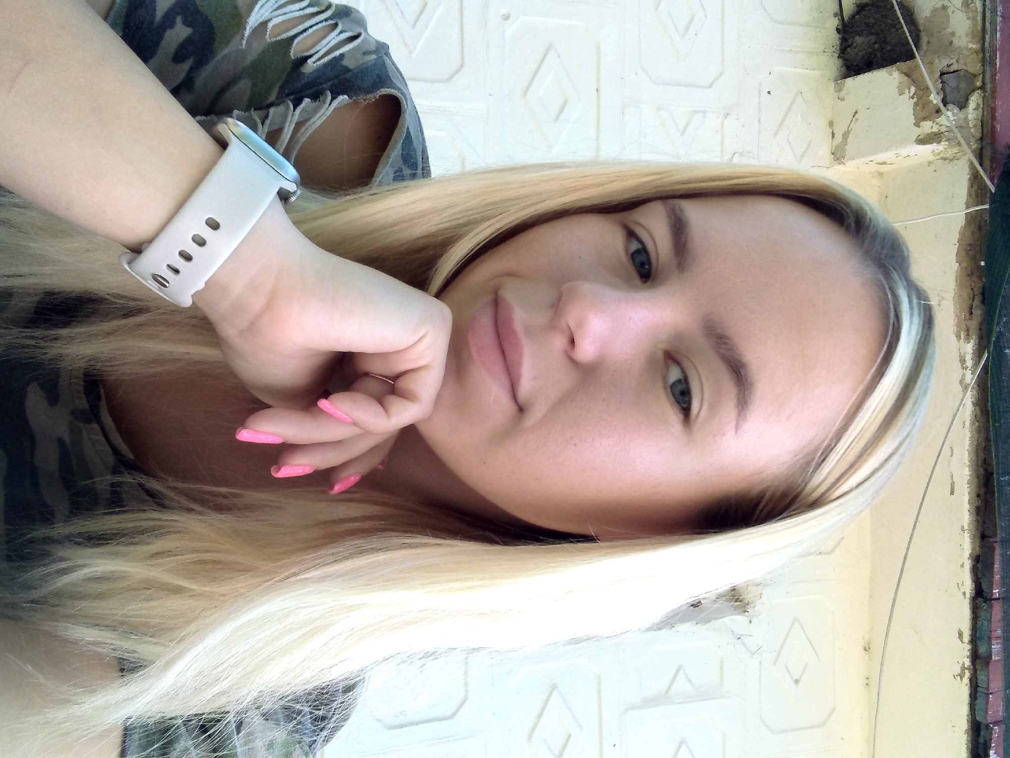 A young woman with blonde hair and blue eyes is pictured from the chest up, resting her chin on her hand. She is wearing a camouflage shirt and a white smartwatch on her left wrist. Her fingernails are painted bright pink. She is leaning against a white textured wall, with a weathered, peeling ceiling and a dark, possibly green, awning visible above. There is a wasp nest attached to the upper left of the image. The scene appears to be outdoors, possibly on a balcony or under a porch in the city of Radisne, Ukraine.