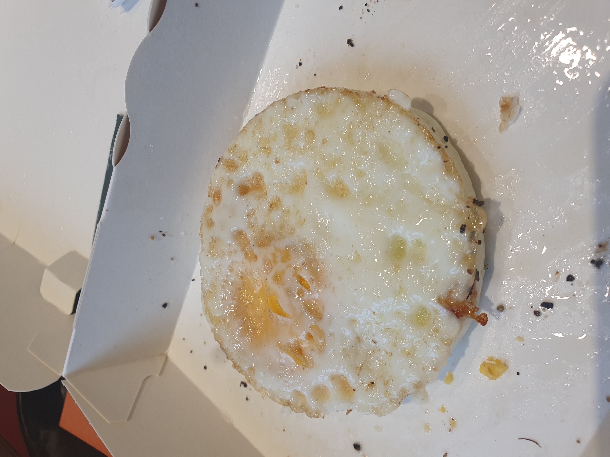 A fried egg is presented in a white cardboard container, likely a takeout box, on a white surface. The egg is cooked sunny-side up, with the yolk slightly broken and visible within the white. The white of the egg is fully cooked, with some crispy, browned edges. Scattered around the egg within the container are small dark specks, possibly pepper or crumbs. The container appears to be slightly greasy, with some moisture visible on the white surface beneath and around the egg. The lighting is bright, indicating an indoor setting. The image is cropped to focus on the egg and its immediate surroundings. The location is Samara, Russia.