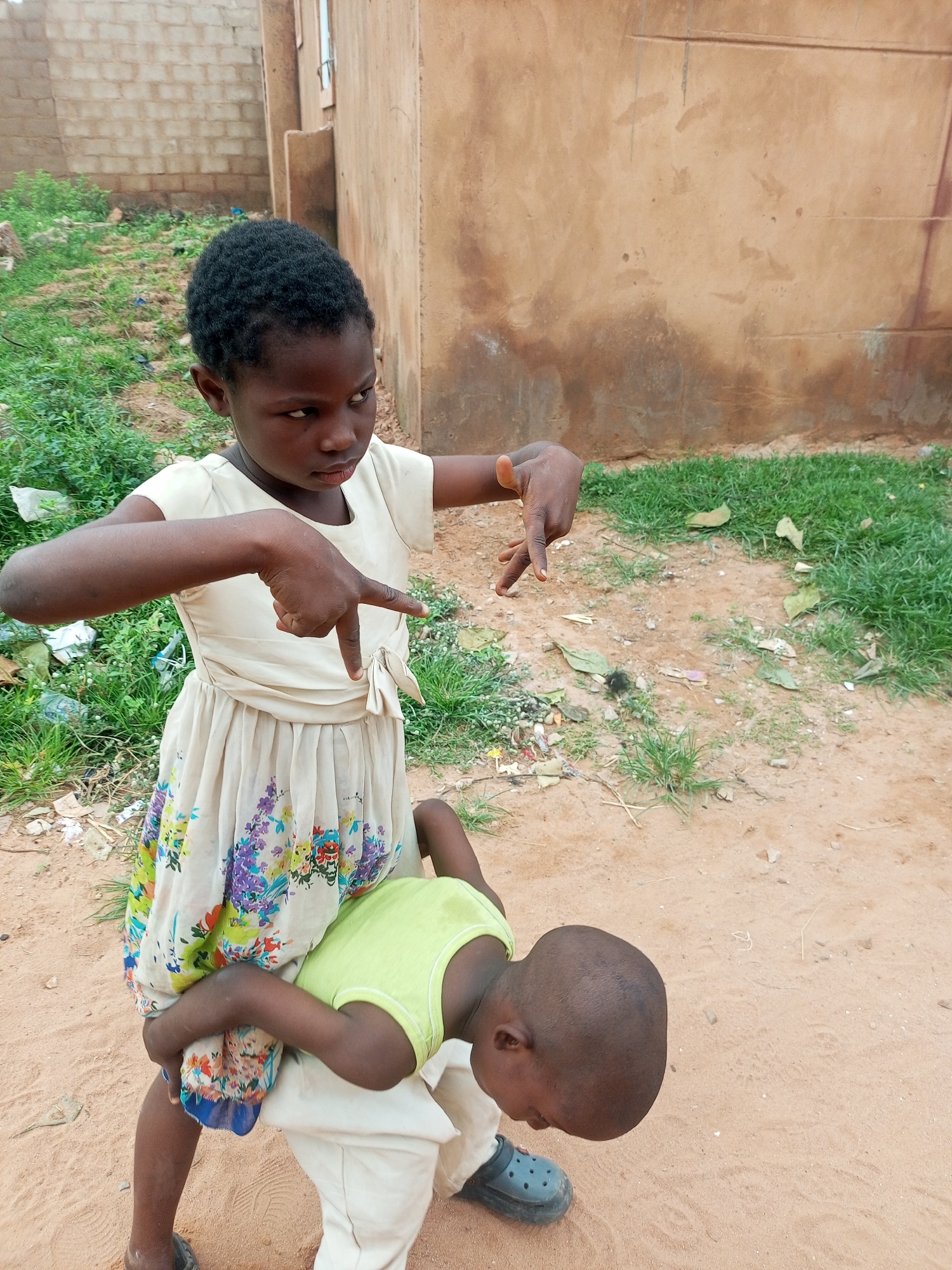 In a dusty outdoor setting in Tempure, Nigeria, a young Black girl stands holding a younger Black boy in a playful embrace. The girl, wearing a cream-colored dress with a floral pattern at the hem and tied at the waist, gestures with her hands. The boy, dressed in a light green t-shirt and off-white pants, is bent over with his head down. He wears dark blue Crocs. The background features a textured, tan-colored wall of a building and a patch of green grass with some scattered debris, suggesting a modest urban environment.