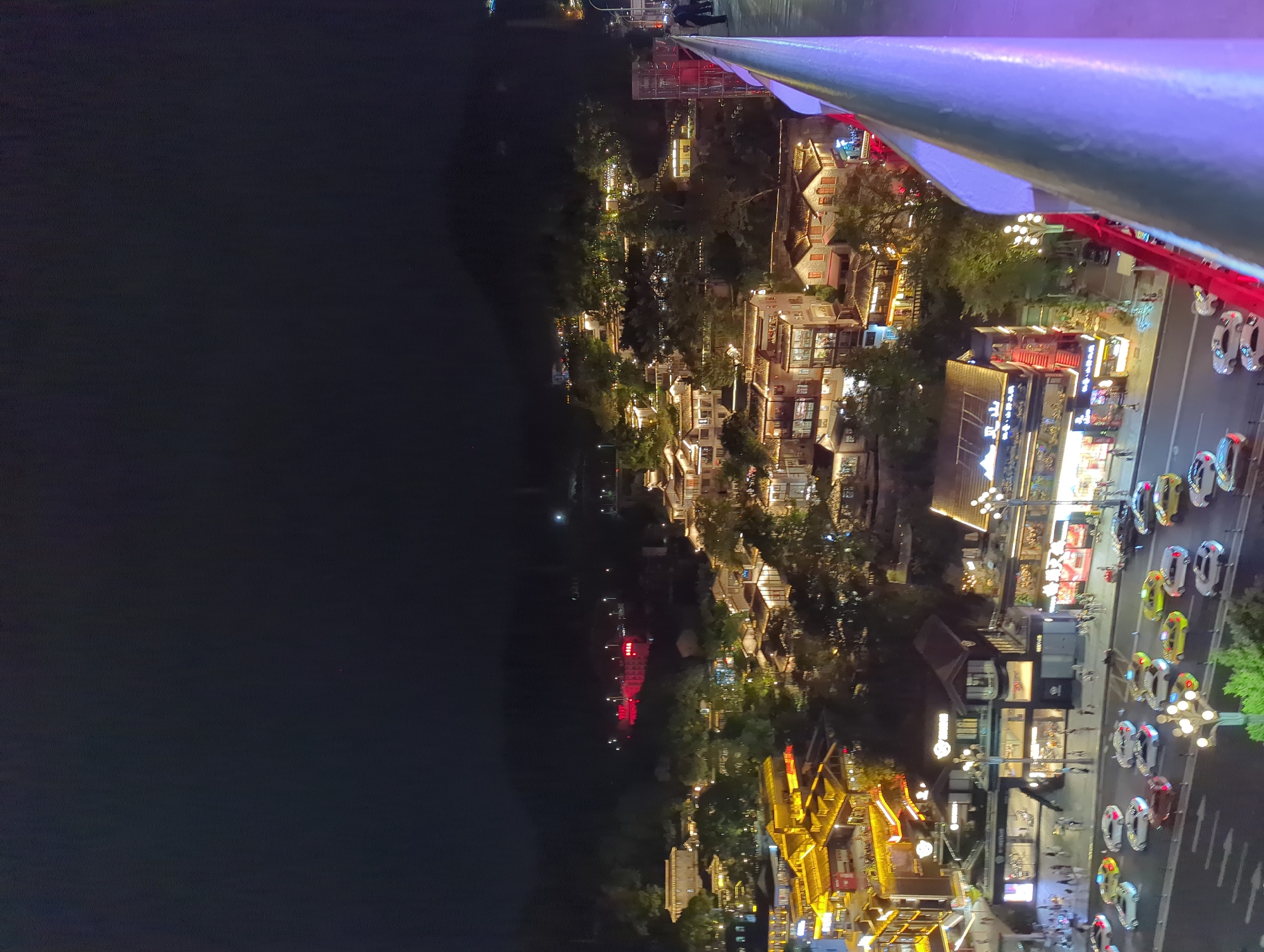 The image shows a nighttime aerial view of a densely populated urban area, likely a historic or tourist district, in Yuzhong District, China. The scene is characterized by numerous traditional-style buildings with illuminated windows and roofs, creating a warm glow against the dark sky. Many of these buildings are clustered together, with some appearing to be shops or restaurants, indicated by storefront signs and open entrances.
In the foreground, a modern pedestrian walkway or bridge railing, illuminated with purple and blue lights, runs along the right side of the frame. Below the buildings, a street is visible, with several cars driving and parked. The cars are a mix of white, yellow, and silver, suggesting common vehicle colors in the region. Pedestrians are also sparsely visible on the sidewalks and near the entrances of the buildings.
The background features a dark, hilly landscape dotted with more illuminated structures, indicating the city extends into the surrounding terrain. The overall atmosphere is one of a vibrant, active nighttime cityscape, with a blend of traditional architecture and modern illumination.