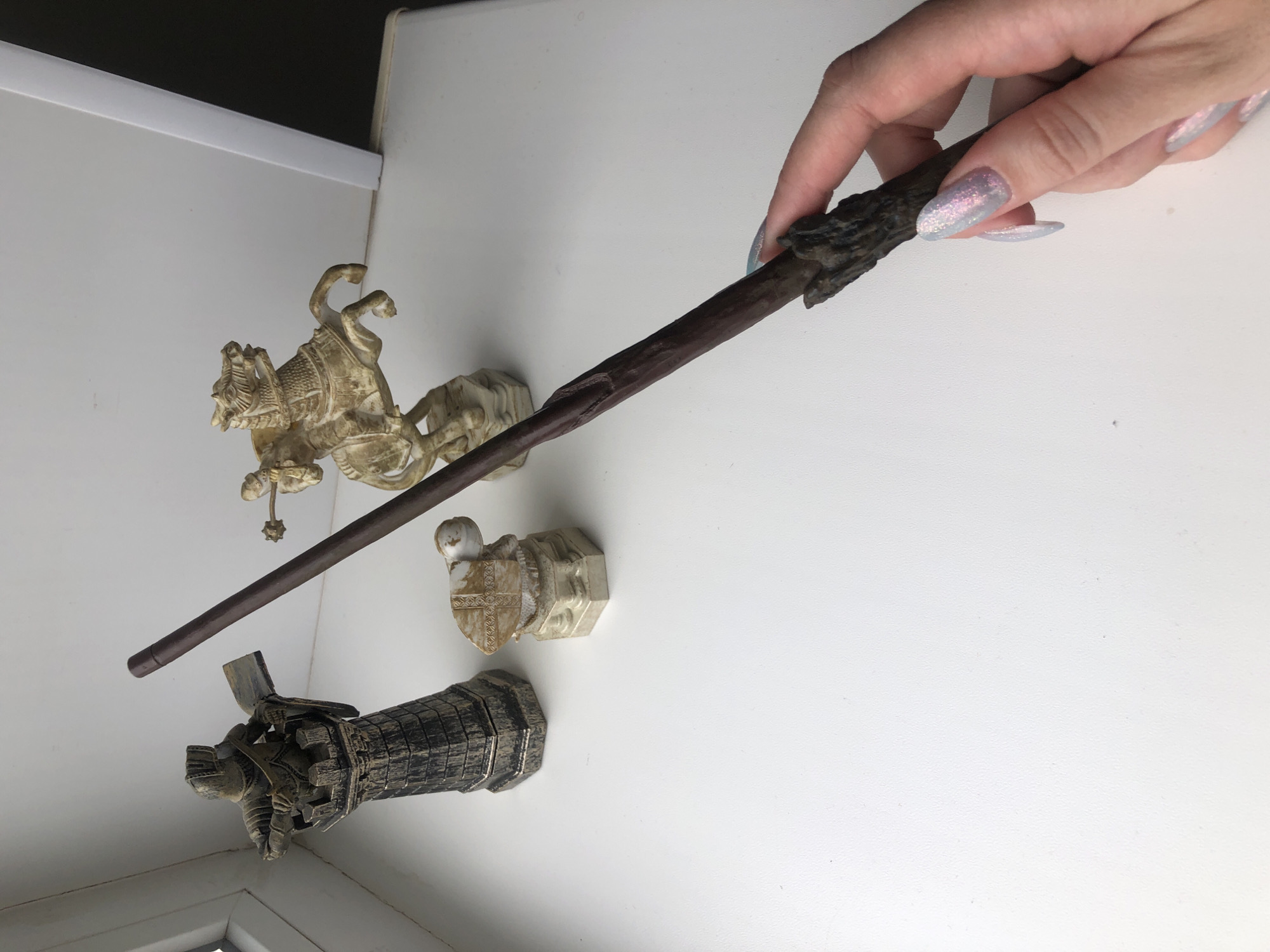 A hand with glittery fingernails holds a brown wand over a white surface. Three small figurines are positioned on the surface: a horse and rider, a shield emblem, and a section of a wall or structure with a miniature figure on top. The objects appear to be resting on a surface near a window.