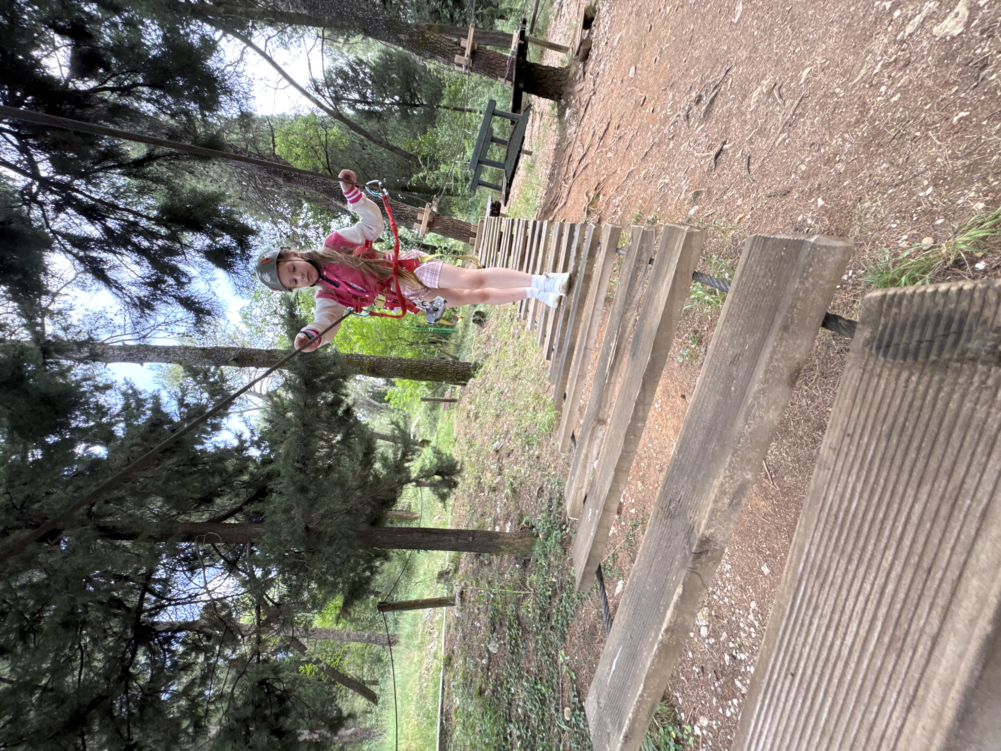 A young girl is navigating a high ropes course in a forest environment, wearing a helmet and safety harness, gripping onto safety cables, and walking across wooden planks. The location is likely an adventure park, given the presence of the ropes course equipment, and it is situated in a wooded area near Podgorica, Montenegro. A bench is also visible in the background. The weather appears to be sunny and dry.