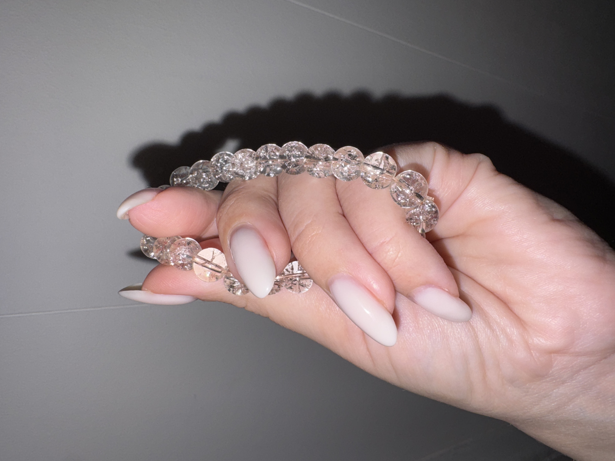 A right hand holds a clear crystal beaded bracelet against a pale gray background. The hand is fair-skinned with long, almond-shaped nails painted a light, creamy color. The beads of the bracelet are transparent and faceted, creating a sparkling effect. A blurry shadow of the bracelet is cast behind it on the wall. The location in Chaikovsky, Russia, is not discernible from the image.