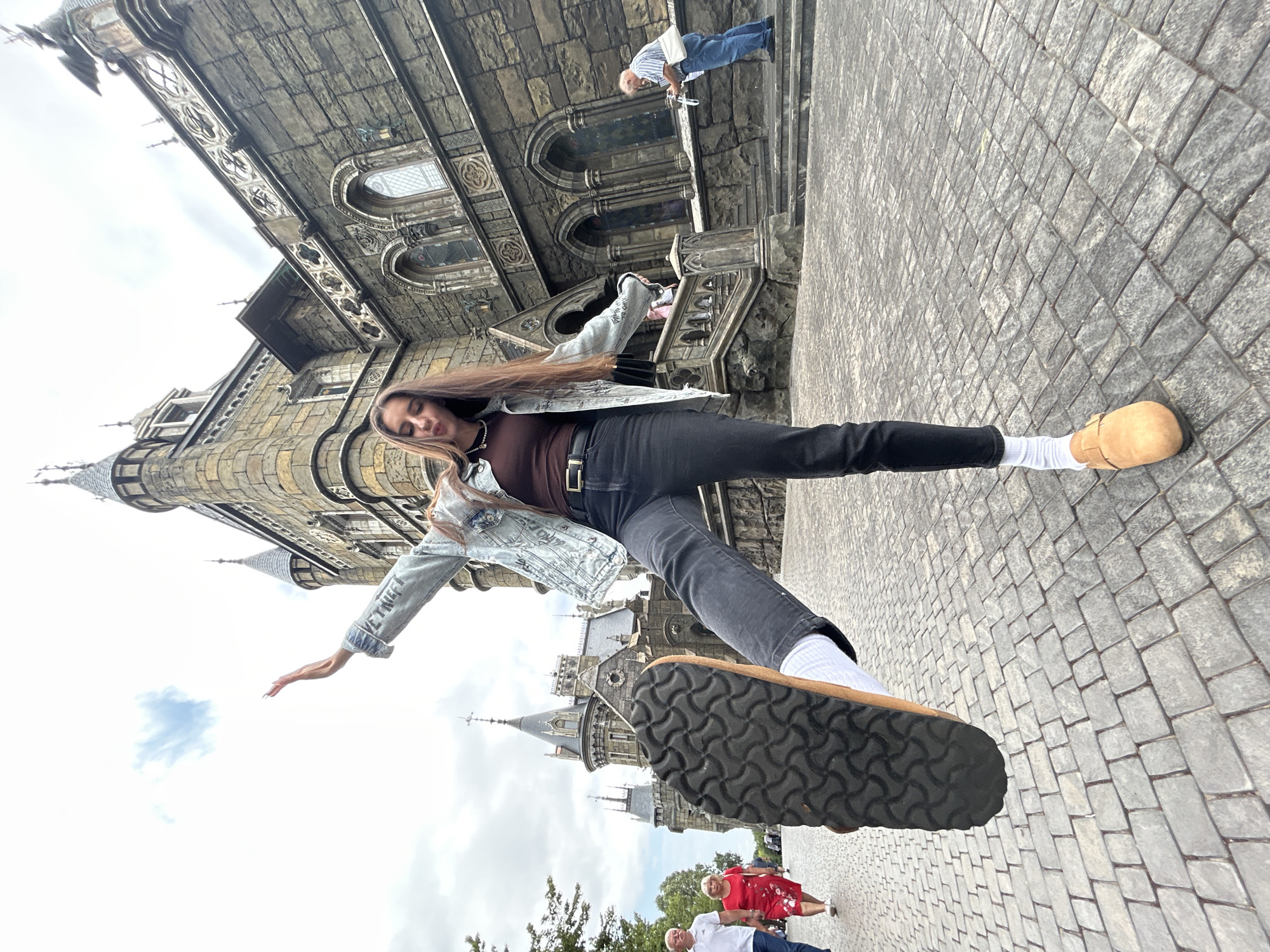 In the city of Khryashchevka, Russia, a young woman stands in front of a stone castle-like building. She is wearing a denim jacket, a brown top, black jeans, white socks, and light brown clogs. Her right leg is extended forward, and her right foot is raised, showing the sole of her shoe. She has long, dark brown hair and is looking down. Her left arm is raised, with her hand extended upwards.
Behind her, the castle has multiple towers and intricate stone carvings. The ground is paved with grey cobblestones. In the background, a couple in red and white clothing walks away from the camera. Further in the distance, an elderly man in a striped shirt and blue jeans stands on steps, holding a white object. The sky is cloudy.