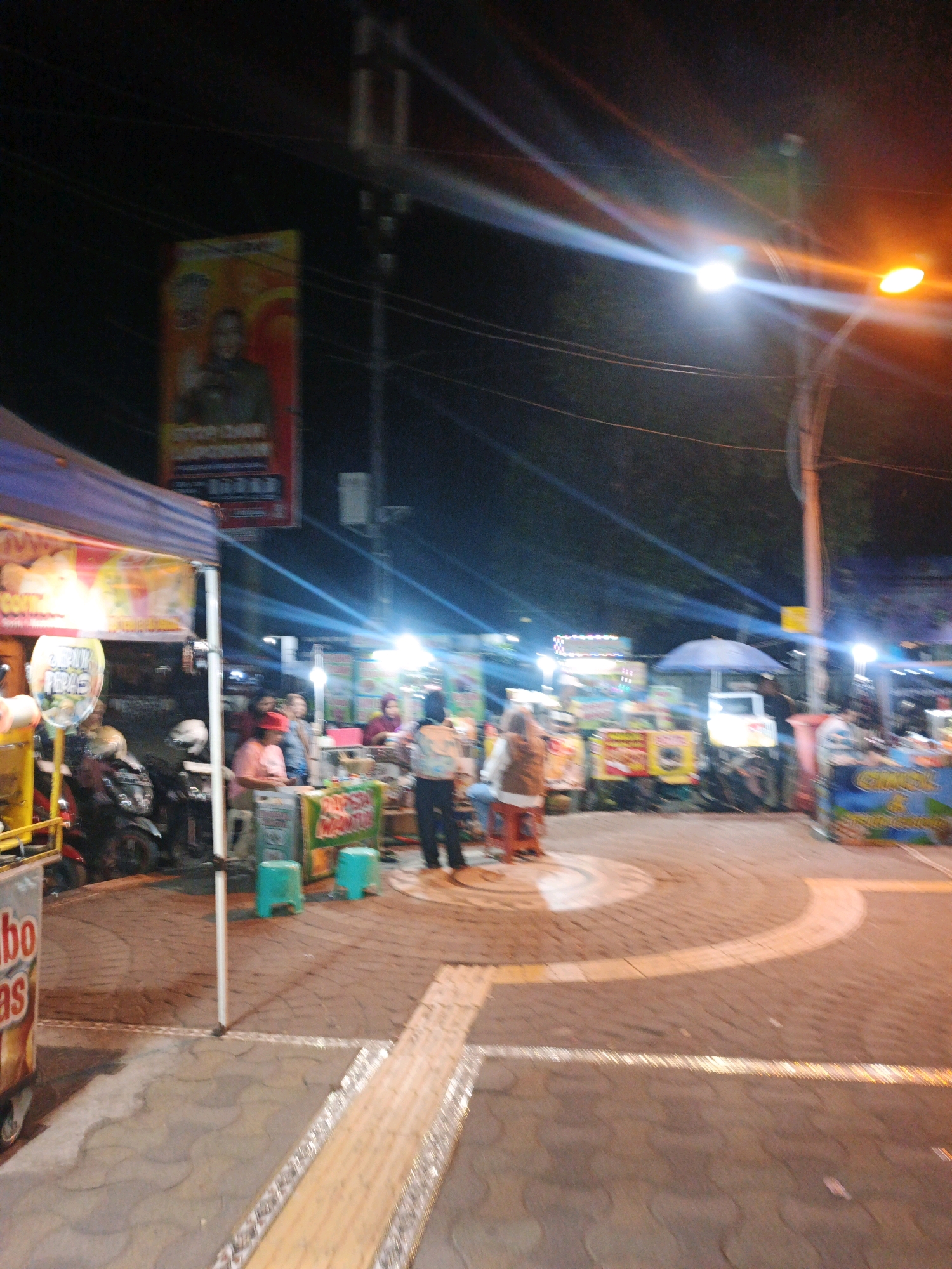 The image depicts a nighttime street scene in Kauman, Indonesia, likely a market or food stall area. Several food vendors are set up, indicated by colorful stalls and signage. In the foreground, to the left, a food cart with "bo as" visible on its side is present. Behind it, a blue tent covers part of a stall with some motorcycles parked nearby.
Further back, the scene opens up to more stalls. One stall has a blue umbrella, and a white vehicle is parked behind it. People are visible at the stalls, interacting with vendors or customers. A tall pole with streetlights illuminates the area, casting bright light and lens flares across the image. The ground is paved with patterned bricks and features a distinctive curved white and yellow marking. A large banner with a portrait is hanging from a pole on the left side of the image. The overall atmosphere is bustling and lively, typical of a night market.