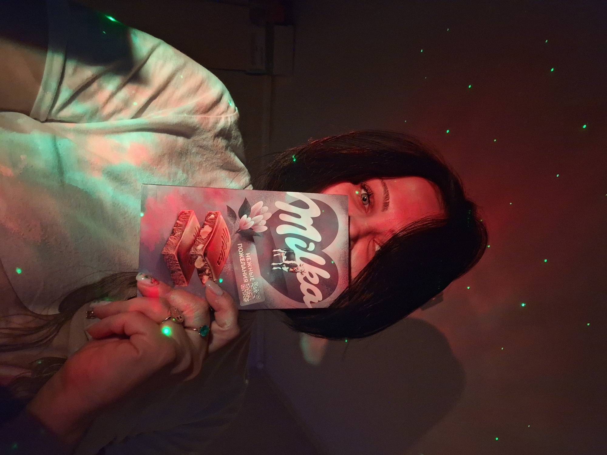 An indoor, close-up shot captures a young woman holding a Milka chocolate bar, which partially obscures her face, revealing only one eye looking towards the camera. She has dark hair and light-colored nails, with several rings visible on her fingers, including one with a prominent green stone.
The setting is dimly lit, with dramatic red and green light projections creating a dynamic, almost festive atmosphere. Small, bright green dots are scattered across the dark background, resembling laser lights. A wash of red light illuminates parts of her face and the chocolate bar, while green light touches her clothing. This lighting suggests an evening or nighttime scene.
The chocolate bar is clearly a Milka brand product, featuring the "Milka" logo and a cow icon. Cyrillic text on the packaging reads "НЕЖНЫЕ ПОЖЕЛАНИЯ," which translates to "TENDER WISHES," along with a QR code. The image conveys a casual, relaxed, and perhaps playful moment, given the pose and lighting. The location context is Moscow, Russia.