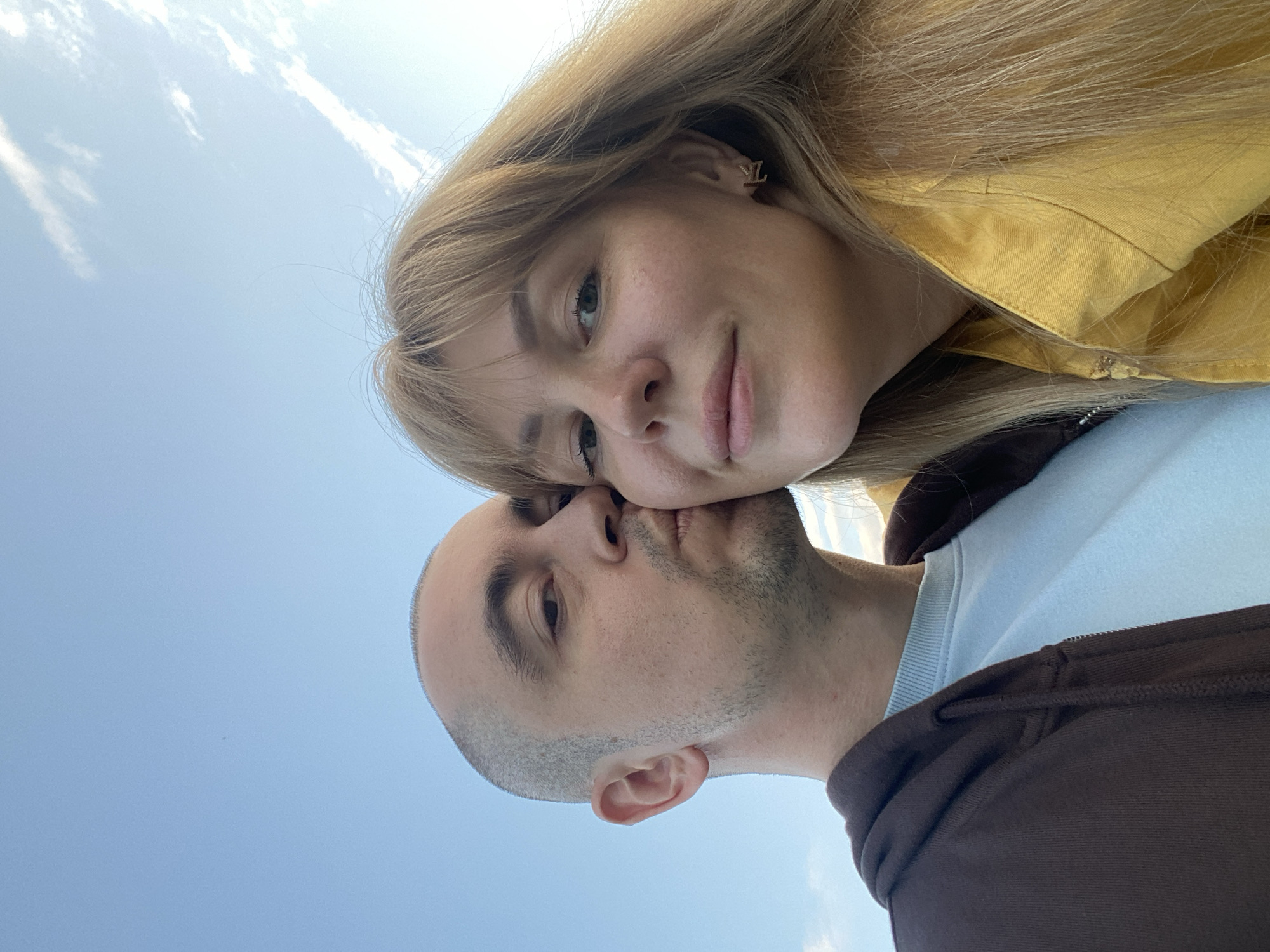 This is a selfie of a young couple outdoors, with a bright blue sky and scattered white clouds filling the background. The couple appears to be in an affectionate pose, with their heads close together. The woman, on the left, has blonde hair and is wearing a yellow jacket. She is looking towards the camera with a gentle smile. Her ear is visible, adorned with a small, letter "L" shaped earring. The man, on the right, is balding and has stubble on his face. He is wearing a light blue t-shirt underneath a brown hooded jacket. His eyes are closed as he leans in towards the woman, suggesting intimacy and closeness. The lighting indicates it is daytime, likely afternoon, given the brightness of the sky. There are no discernible location cues in the image itself, but the provided context places the scene in Yekaterinburg, Russia. The overall mood conveyed is one of love and happiness.