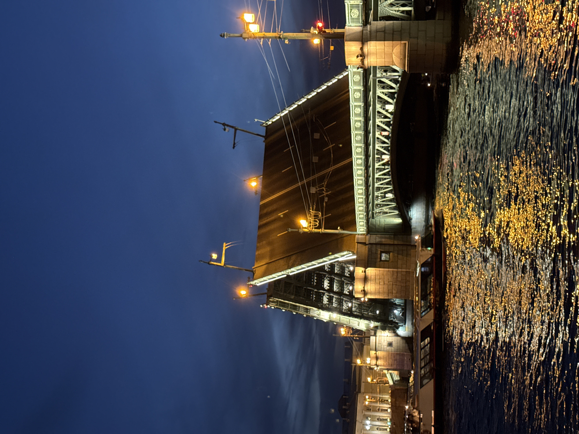 The image depicts the Palace Bridge (Dvortsovy Most) in Saint Petersburg, Russia, during nighttime. The drawbridge is in its open position, with its two central bascule leaves raised upwards at a significant angle, illuminated by warm yellow lights outlining their structure and sides. The sky above is a dark, deep blue with subtle cloud formations.
The Neva River flows beneath the bridge. Its surface is dark, reflecting numerous shimmering points of golden-yellow light from the bridge and adjacent structures, creating a ripple effect. On the left side of the frame, a long, low passenger boat with illuminated windows is visible on the river, positioned near the riverbank. Another boat or a part of one might be partially visible further right on the water.
The bridge's fixed spans, constructed with metal trusswork, are visible below the raised leaves and to the right. The bridge rests on robust stone piers. On the right, a tall, multi-armed street lamp post with multiple yellow lights and visible power lines extends upwards. Buildings with classical architecture, also illuminated, line the left riverbank in the background, including a structure with columns. Additional streetlights are visible on both the bridge and the surrounding embankments. No discernible individuals are visible within the frame.