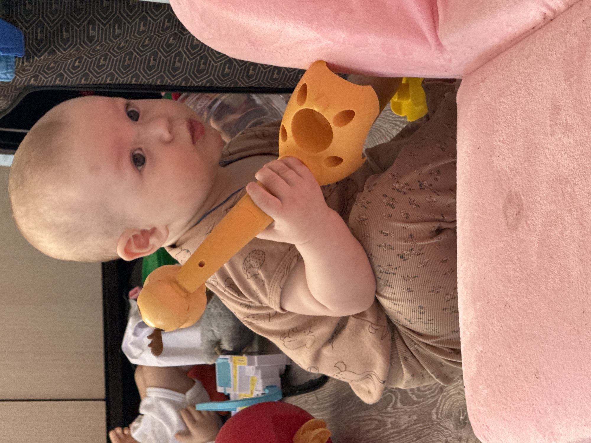 A baby is holding a toy in front of a pink couch. There are other toys visible around the baby. The location is unknown, as the city of Кипчак, Russia is not mentioned in the image context.