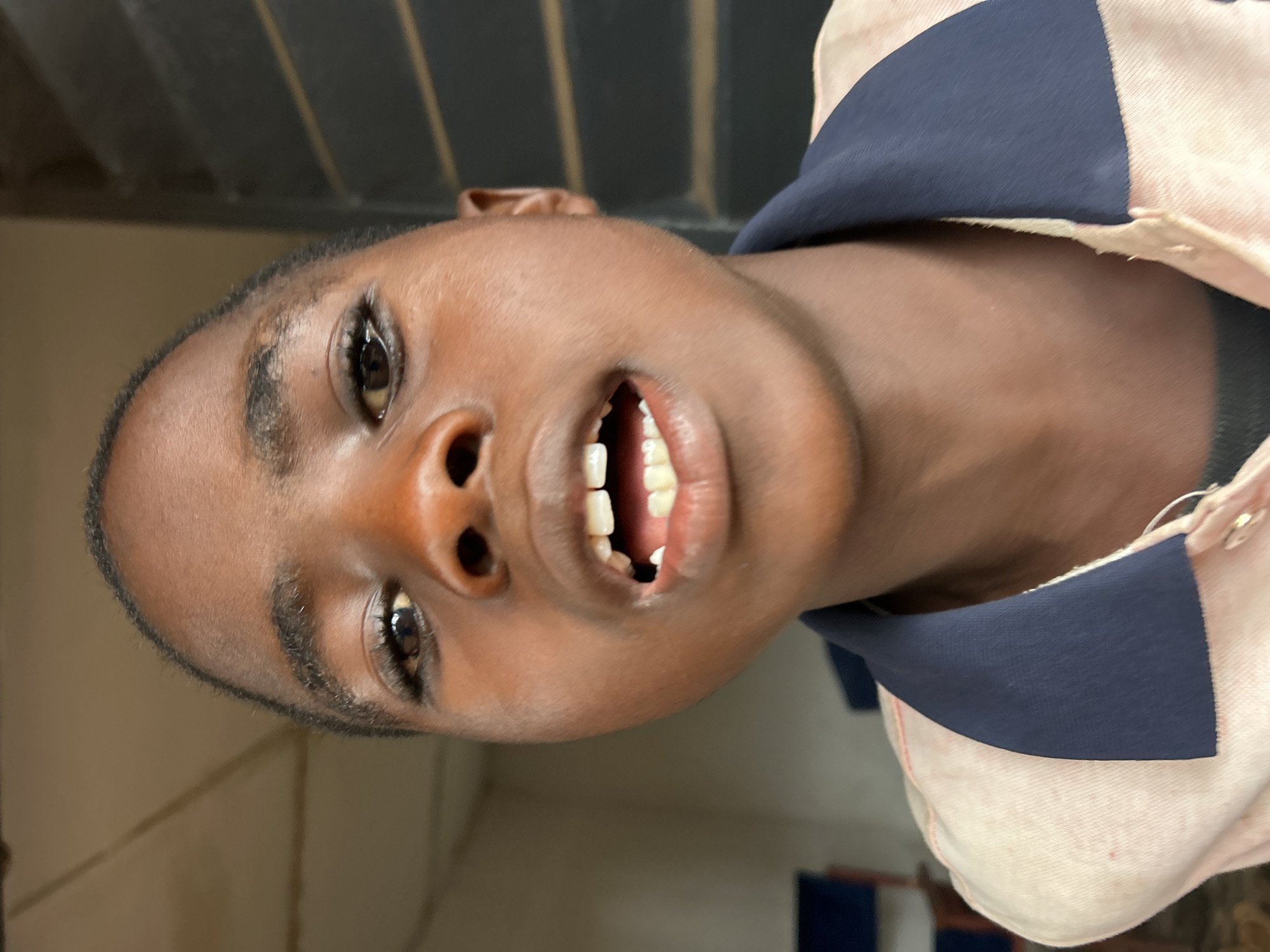 A close-up, angled view depicts a person with dark skin and short, dark hair, wearing a pale pink shirt with a navy blue collar. The person's mouth is open, revealing their teeth. The location is unknown.