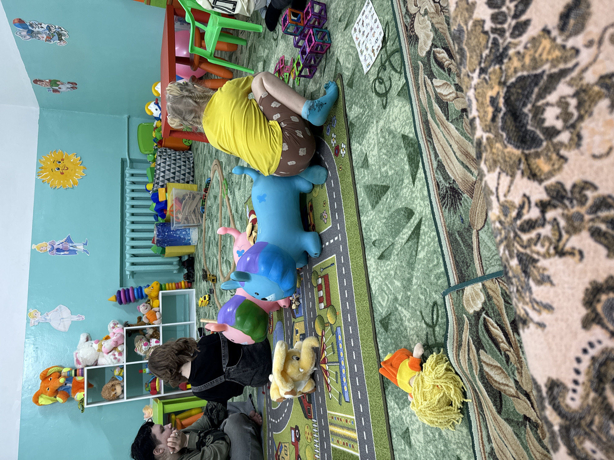 A photo depicts a brightly colored playroom interior with various people and objects. A child is riding a blue toy animal on a carpet with a road pattern, while another individual is kneeling nearby. A shelf filled with stuffed animals sits against the blue-painted wall, adorned with cartoon character decals. The room features a small red table with colorful chairs. There's a large floral-patterned rug partially covering a green rug. The scene is in Anapa, Russia.