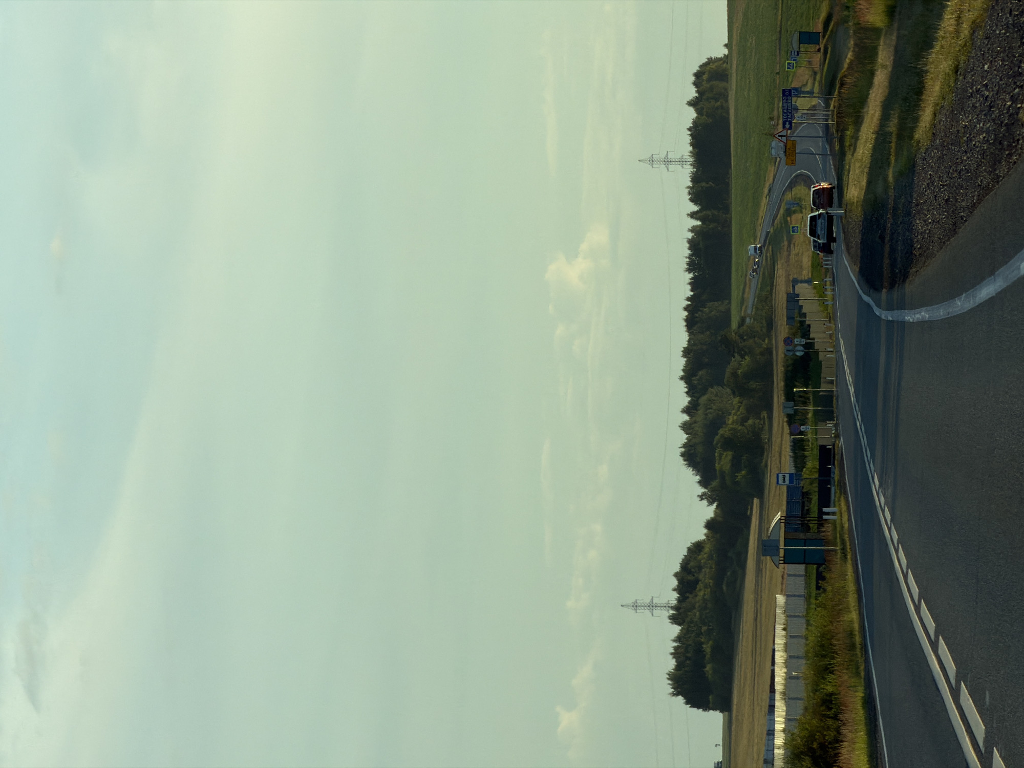 The image depicts a roadside scene in the vicinity of Togashevo, Russia, likely during daylight hours given the illumination. A paved road curves through a landscape of rolling hills and vegetation. Several vehicles, including cars and what appears to be a van, are visible on the road, some moving and others parked. The presence of traffic signs, a bus stop shelter, and a white barrier suggests this is a populated area or a route with infrastructure. In the background, trees form a dense line along the horizon, with power lines and towers indicating the presence of electrical infrastructure. The sky is a pale blue with scattered clouds. The overall impression is of a rural or semi-rural setting on a highway.