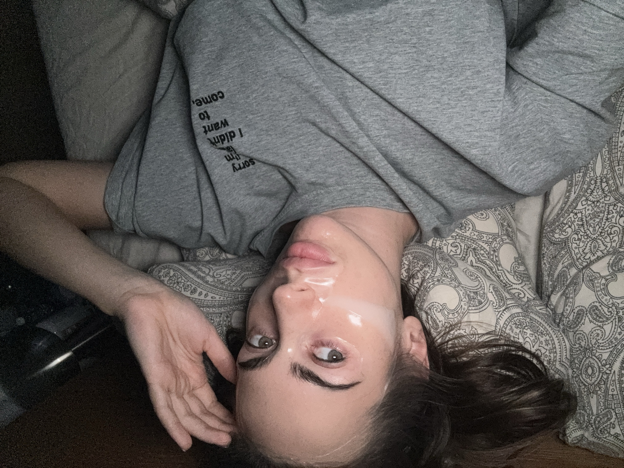 A woman lies on a patterned pillow, wearing a gray t-shirt with the text "sorry I didn't want to come." She has a transparent mask on her face. The setting appears to be indoors, likely a bedroom. There is no indication of the location being in Warsaw, Poland.