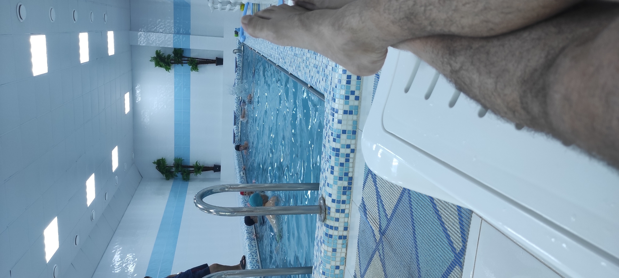 This is a medium-shot, eye-level, outdoor, day-time photo taken in Noviy, Russia. The photo captures a person's legs, which are hairy and tanned, resting on a white lounge chair. The perspective is from the person's point of view, looking towards an indoor swimming pool. The pool is filled with clear blue water and has a tiled edge. Several people, including children wearing swimming caps, are visible in the pool, some are swimming while others are being instructed. There are two artificial palm trees on either side of the pool, and the walls are tiled in white and blue. The ceiling is white and features fluorescent lighting panels. The floor around the pool is tiled and a patterned mat is visible.