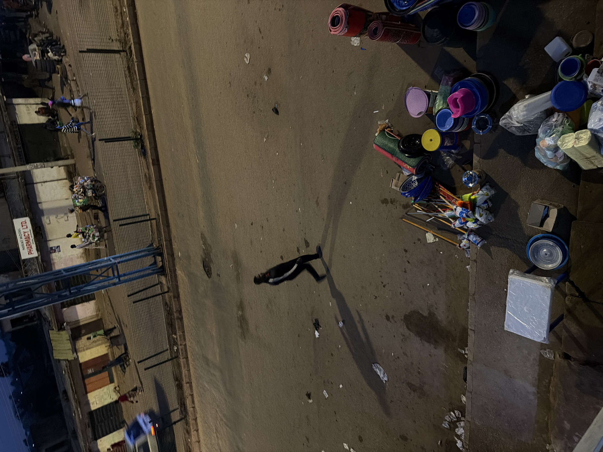 The image depicts a street scene in Bukuru, Nigeria, likely during the late evening or early morning due to the low light conditions. The foreground shows a collection of miscellaneous items, including stacked plastic basins of various colors (blue, pink, purple), rolled mats or carpets, brooms, and some plastic bags of goods. A small white table covered in plastic wrap is also visible.
In the mid-ground, a lone figure dressed in dark clothing with white stripes down the sleeves and legs walks across the asphalt road. The figure's shadow is cast long across the pavement. The road surface is littered with small pieces of debris.
The background features a row of buildings that appear to be shops or market stalls, some with closed shutters. A vehicle, possibly a car, is visible on the left side of the road, appearing somewhat blurred, suggesting movement. Several other figures are present in the background, some near the buildings and others near a cart or bicycle, possibly engaged in activities related to transportation or commerce. A prominent blue metal structure, possibly a utility pole or part of an overhead sign, dominates the right side of the background. A sign reading "NERAT MODEL PHARMACY LTD." is visible on one of the buildings. The overall impression is that of a busy commercial area at a quiet time of day.