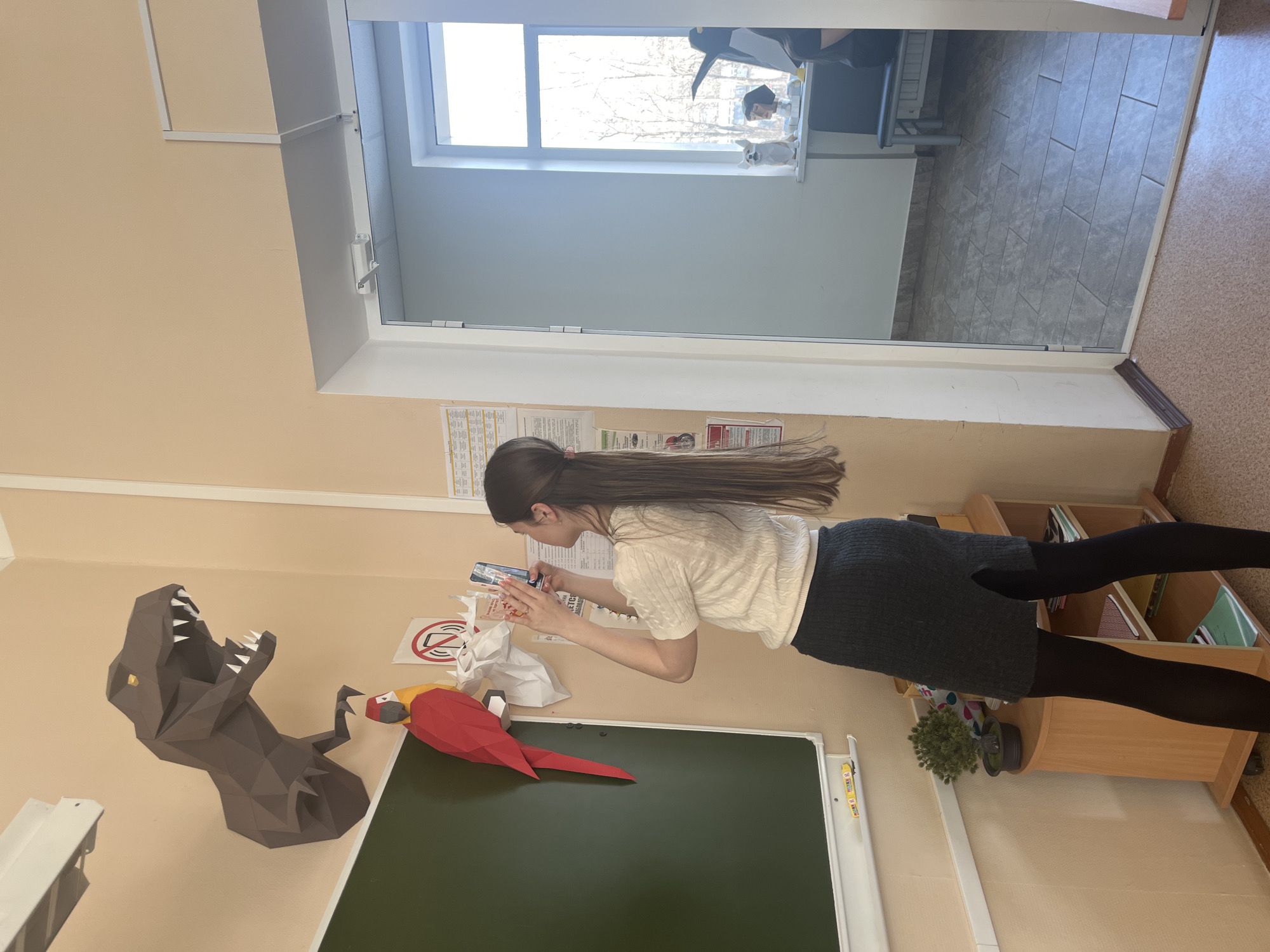 A young woman with long, dark brown hair, wearing a light-colored textured top, a dark grey skirt, and black tights, stands in what appears to be a classroom or educational facility in Chaikovsky, Russia. She is focused on using her smartphone, holding it with both hands, seemingly taking a picture or video of the wall decorations.
The room features light beige walls and a dark green blackboard. On the wall above the blackboard are two large, polygonal papercraft figures: a grey T-Rex head with its mouth open, showcasing white teeth, and a red and yellow macaw. Near the woman's hands, a white sign with a red circle and diagonal line (a typical "no" symbol) is visible, along with another white paper sign that reads "НЕТ СЛАДКОМУ" ("NO SWEETS") and "ПЕЧЕНЬКАМ" ("COOKIES"), followed by "ДЕТСКАЯ РАБОТА." ("CHILDREN'S WORK."). Further back on the wall, there's a larger white document, likely a schedule or information sheet.
To the right, a wooden bookshelf or cabinet holds various books, binders, and a small potted plant. An open doorway reveals a tiled floor in an adjacent room. A large window provides natural light, showing bare trees outside, suggesting an autumn or winter daytime setting. On the windowsill of the adjacent room, several figurines, including a black penguin and a small white dog, are visible. The floor in the foreground is a light brown or beige, while the floor seen through the doorway is grey tiled. The woman appears engaged in her activity, though her face is not visible.