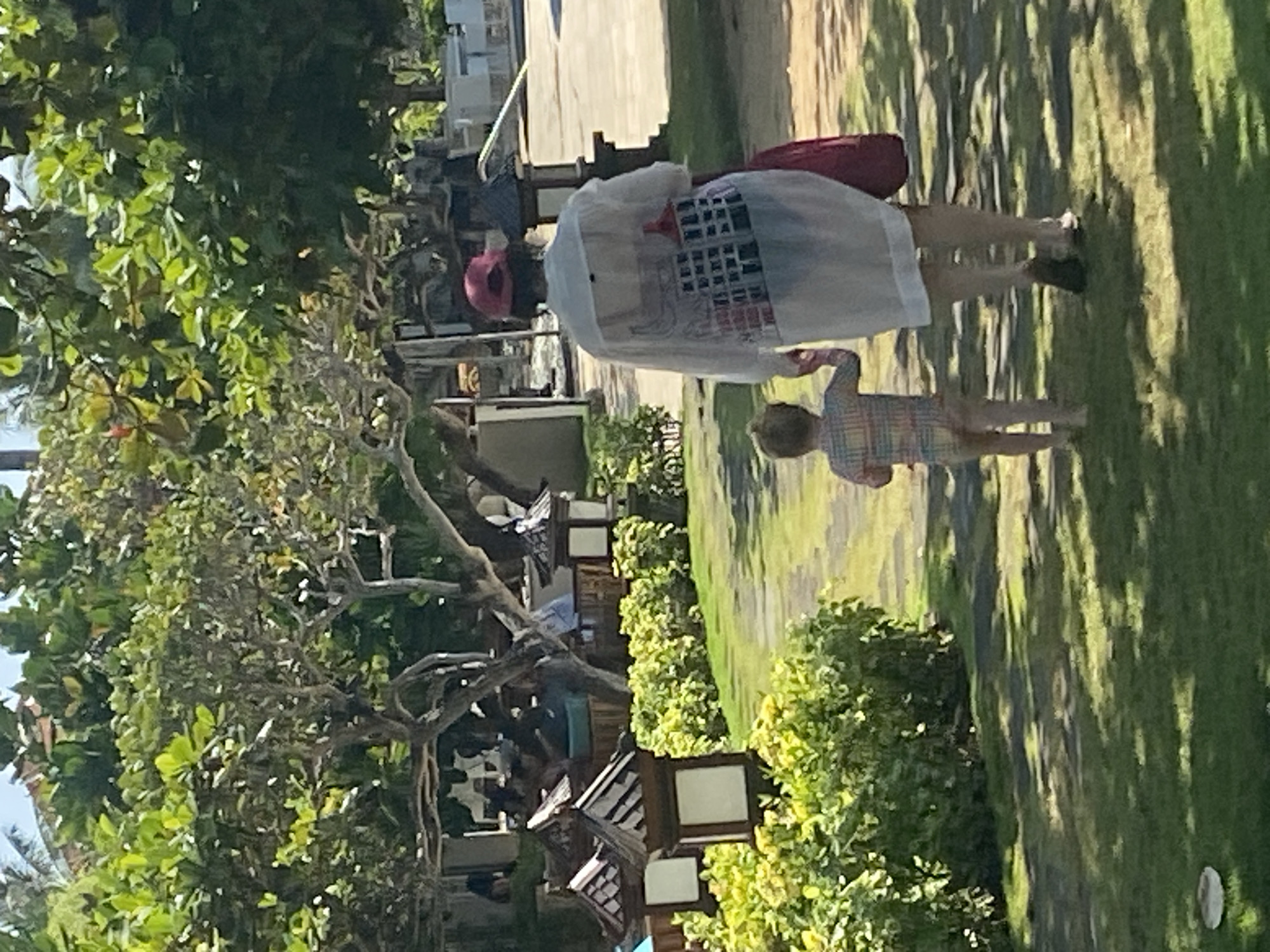 A medium, outdoor shot depicts a woman and child on a manicured lawn in Nusa Dua, Indonesia. The woman is walking right, wearing a light-colored cover-up with a graphic design on the back, a dark bag, and dark shoes. The child, in a plaid shirt, appears to be running in the same direction as the woman. Behind them are trees, shrubs, and decorative light fixtures, indicating a well-maintained resort or residential area. A building can be seen in the background.