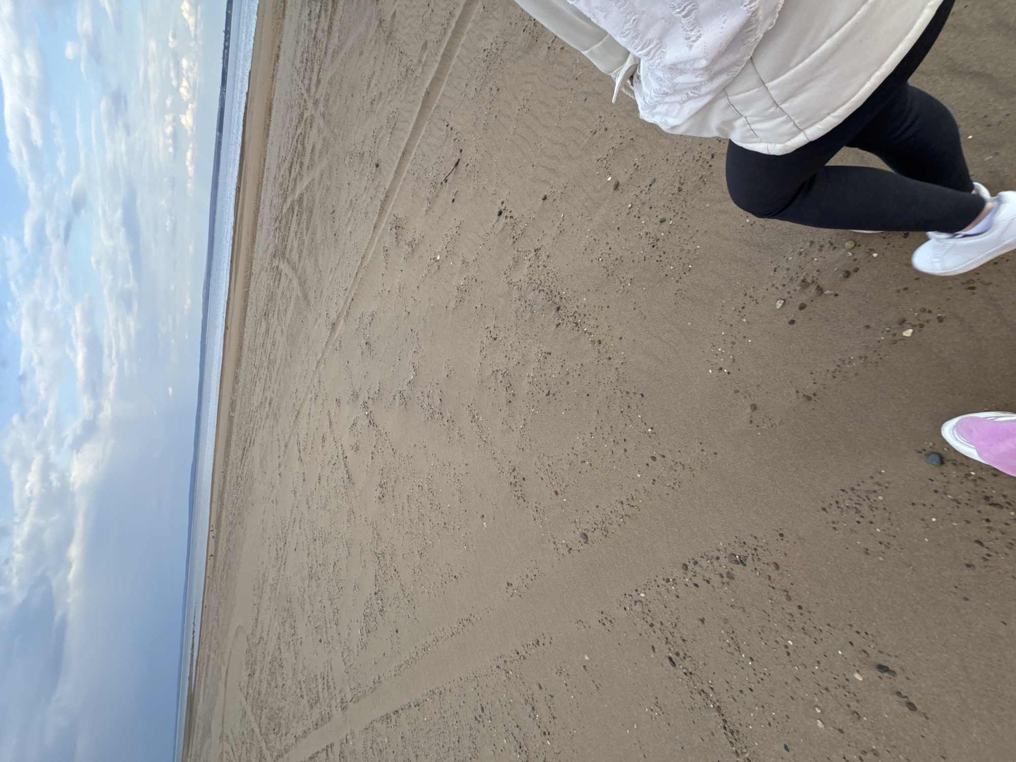 The image shows a beach in Chaikovsky, Russia, during the day. The sky is overcast with patchy clouds. The beach consists of dark sand with small stones. Two people are visible, one wearing black leggings, a white puffer jacket, and white sneakers, and the other wearing pink sneakers. They are walking on the beach. Tire tracks are visible in the sand. The ocean is present in the background.
