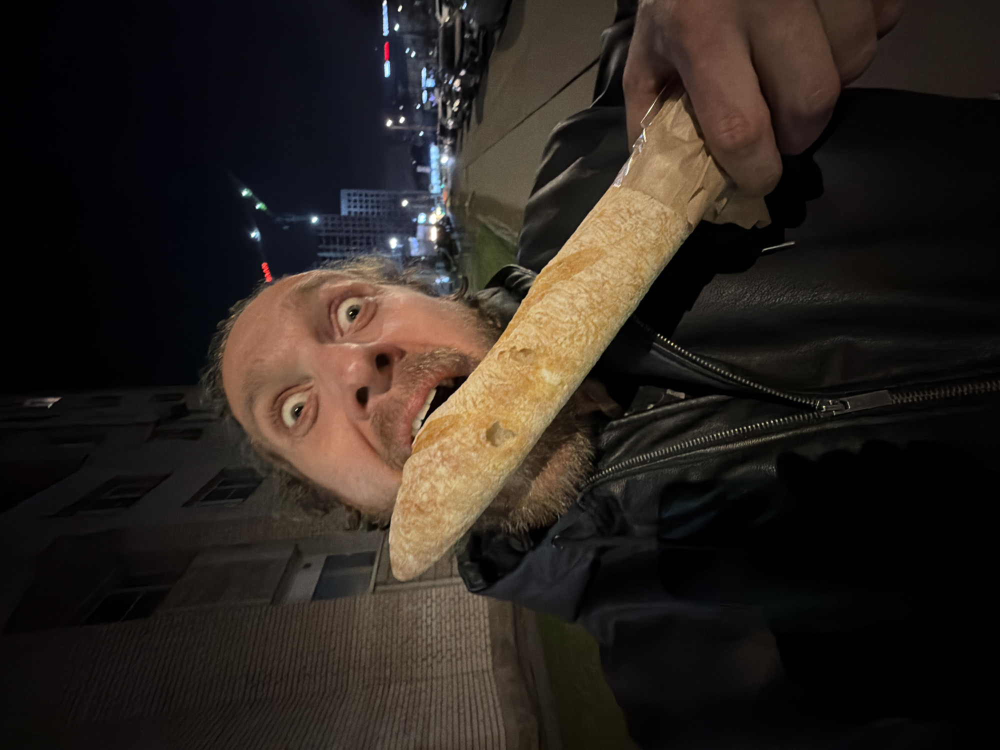 A man is holding a baguette in his mouth, outdoors at night. The man has light skin, curly hair, and a beard. He is wearing a black leather jacket. The baguette is partially wrapped in brown paper. The background includes buildings and streetlights. The scene is likely in Saint Petersburg, Russia, based on the architecture of the buildings in the background.