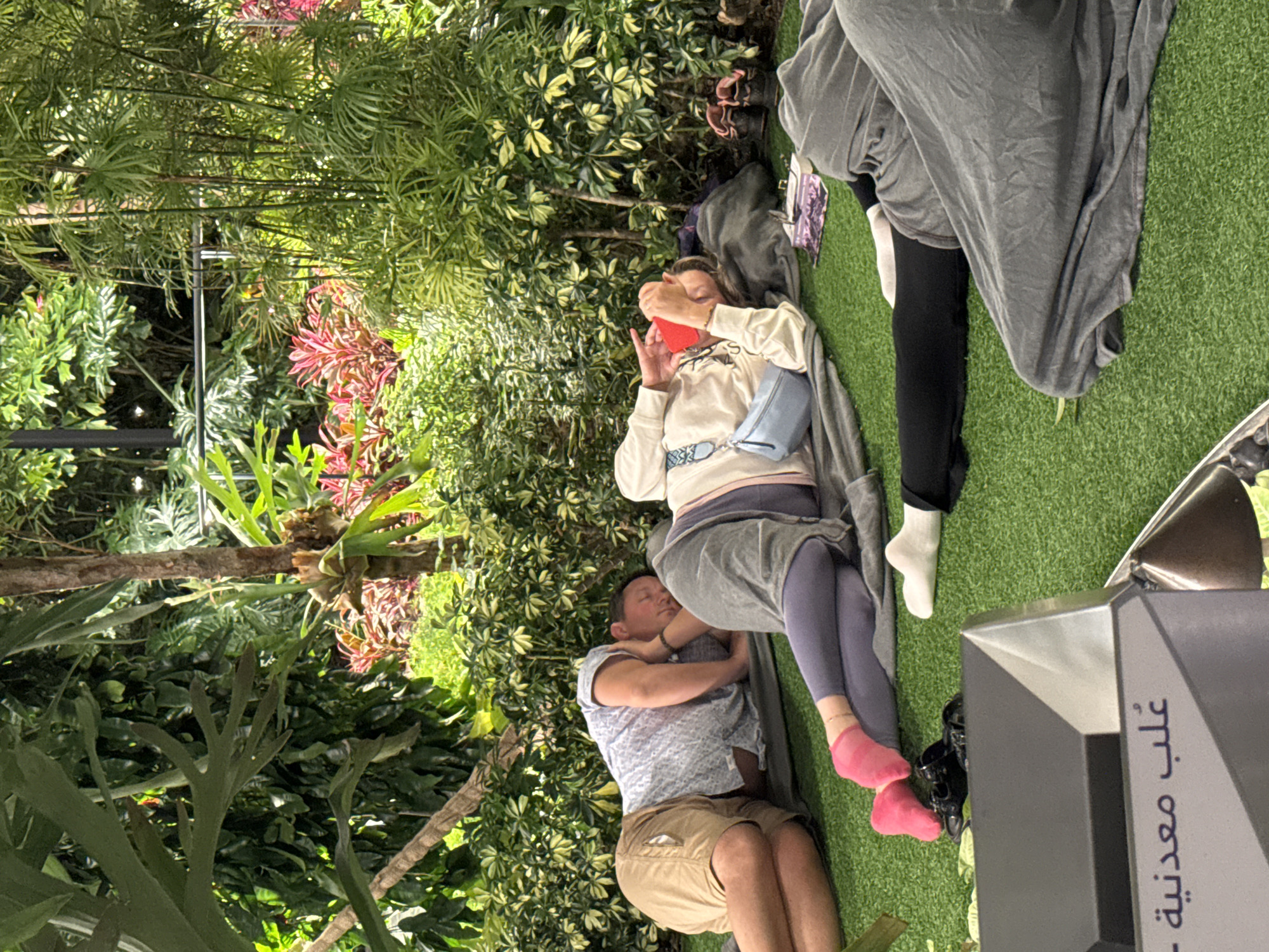 The image depicts a serene scene in a lush, garden-like environment, possibly an indoor or shaded outdoor space within Doha, Qatar. In the foreground, a dark, angular object with Arabic script, likely an informational display or part of an exhibit, is partially visible.
The main focus of the image is on several individuals resting on a bright green, artificial turf surface, all appearing to be asleep or in a relaxed state. To the left, a man is lying on his side, wearing a blue patterned short-sleeved shirt and tan shorts. His right arm is under his head, and his eyes are closed. Next to him, a woman is also lying on her side, facing away from the camera. She is wearing a cream-colored hooded sweatshirt, purple leggings, and bright pink socks. She has a small, light blue crossbody bag slung over her shoulder and is holding a red object, possibly a phone or a small book, near her face. Her eyes are closed, and her head is tilted back. Further to the right, the lower legs and feet of two other individuals are visible, also seemingly resting or sleeping on the turf, covered by grey blankets. One person is wearing white socks, and the other black leggings.
The background is dominated by dense, vibrant greenery, suggesting a botanical garden or a heavily landscaped area. Tall, slender bamboo-like plants are visible to the right, while broader, leafy plants and foliage fill the rest of the background. Red and pink foliage adds pops of color among the green. The overall impression is one of tranquility and repose amidst a natural, verdant setting.