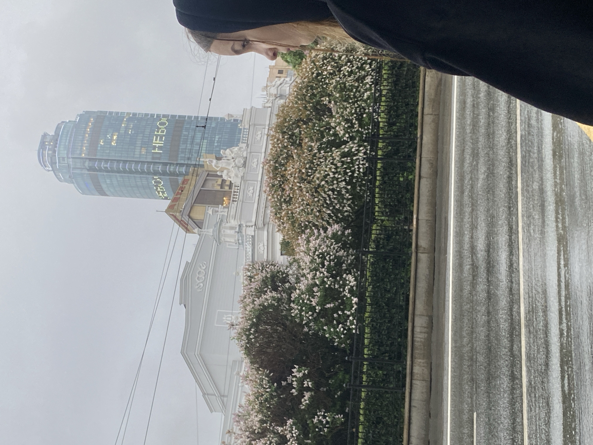The photograph depicts a street scene in Yekaterinburg, Russia, under an overcast sky. A portion of a person's face and hooded garment is visible in the upper portion of the frame. A multi-lane street wet with rain runs vertically along the right side of the frame. A green hedge and a black wrought-iron fence are situated adjacent to the street. Behind the fence are older buildings, including one with statues and architectural details in a light color. In the background, a modern skyscraper displays the word "HELLO" in large, illuminated letters. Electrical wires are strung overhead.