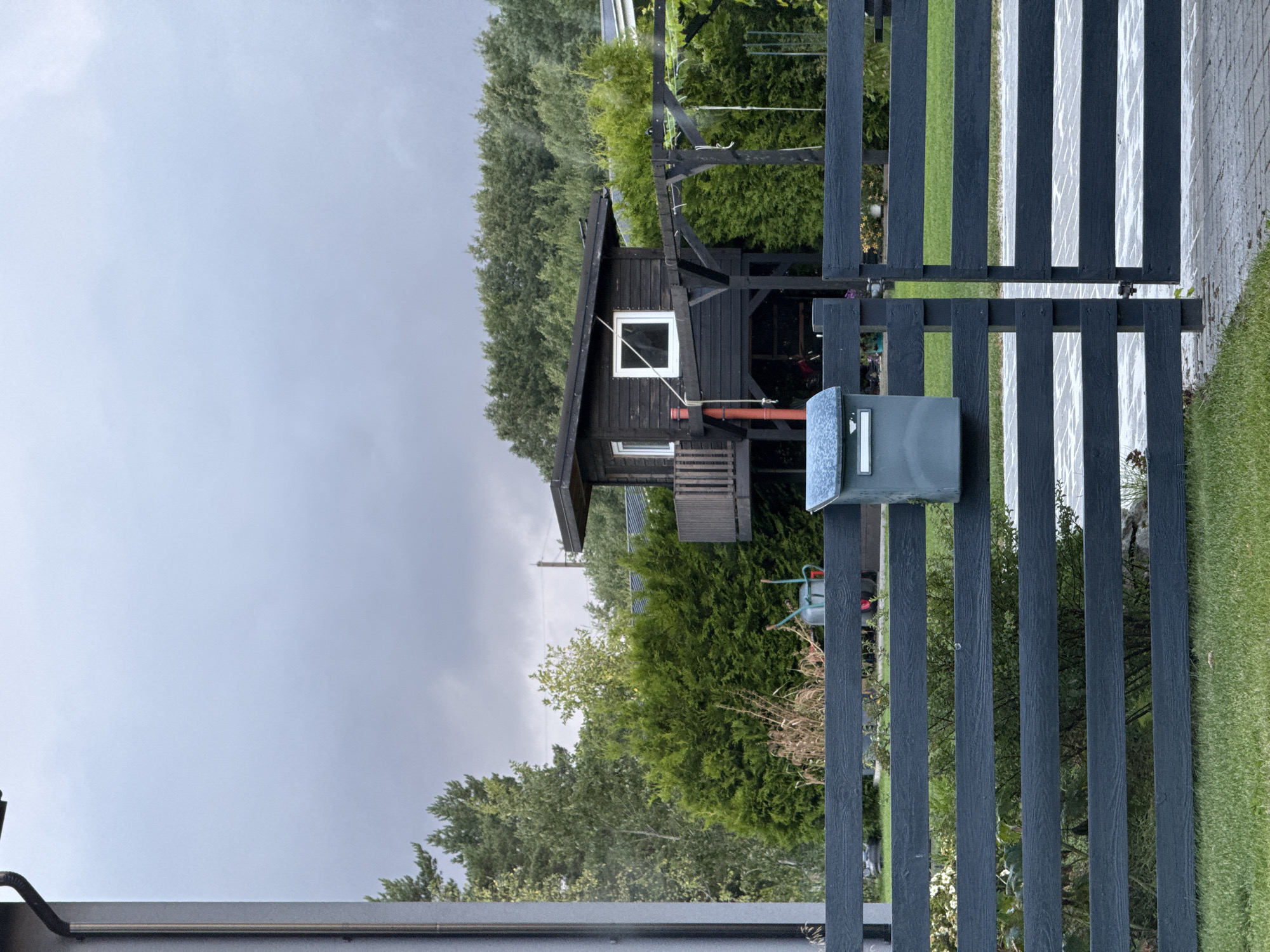 The image depicts a dark grey wooden fence with horizontal slats. A grey mailbox is affixed to the leftmost post of the fence. Beyond the fence, a dark wooden structure, possibly a playhouse or small cabin, is elevated on stilts and features a small balcony and white-framed windows. Dense green coniferous trees and other foliage are visible behind the structure. The sky is overcast with grey clouds. The scene appears to be in a suburban or rural setting, consistent with the Uuesalu area of Estonia. There are no people or animals visible in the image. The ground in the foreground is covered in green grass, and the ground behind the fence appears to be paved with interlocking bricks.