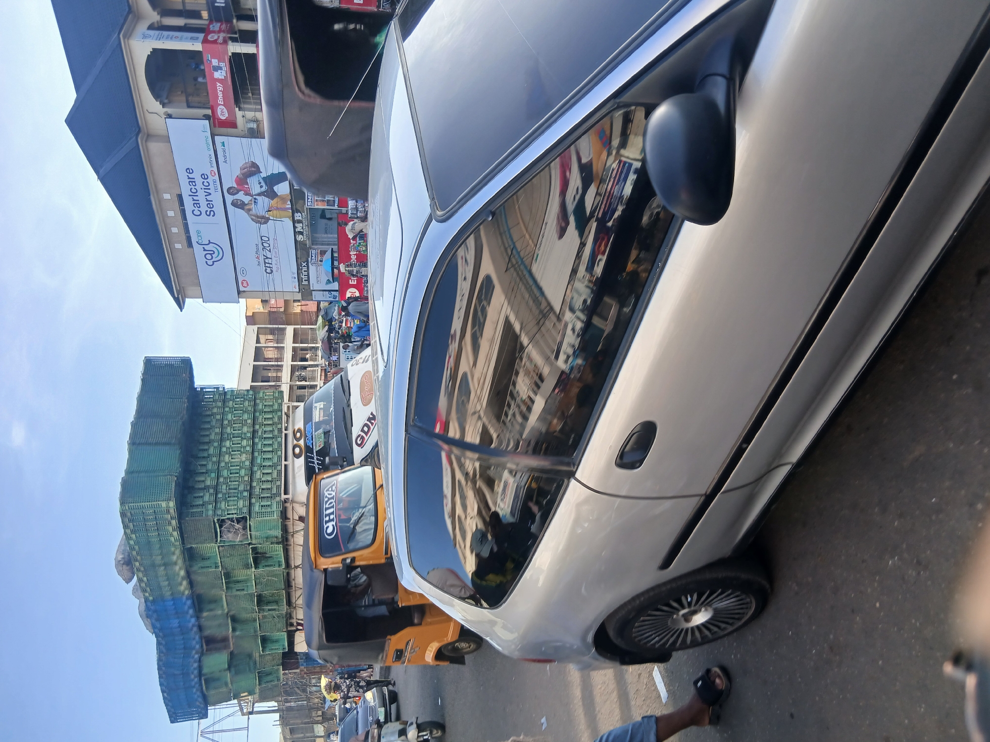 A busy street scene in Maiburiji, Nigeria, is captured during bright daylight with a clear sky. The image focuses on the right side of a silver car, showing its passenger side door, wheel, and the reflection of surrounding buildings in its windows. In the immediate foreground, a person's foot, clad in a black sandal, is visible next to the car.
Behind and to the left of the silver car, an orange three-wheeled vehicle, possibly an auto-rickshaw or "Keke Napep," is present, bearing the word "CHIYA" on its windshield. Further back, part of a larger vehicle, perhaps a bus or truck, shows "GDN" on its side. To the far left, a towering stack of green plastic crates, with some blue crates at the base, is loaded onto an unseen vehicle, indicating the transport of goods.
The background features multi-story commercial buildings adorned with various advertisements and shop signs, suggesting a bustling commercial area. Visible text on these signs includes "Caricare Service," "TECNO," "Infinix," "oralmo," "Android," "itel Ai Phone," "CITY 200," "Pop Fun Ever Strong," "SMB COMMUNICATION," and "itel Energy." Several people are discernible in the background, moving between the vehicles and shops, contributing to the lively atmosphere of a typical market or street day in Nigeria. The overall scene depicts vibrant everyday activity and commerce.