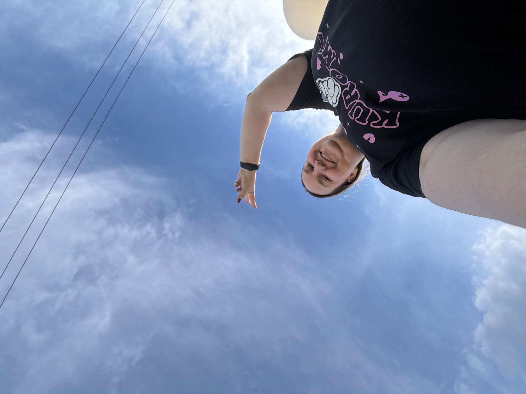 The image shows a person looking down at the camera from a high angle, against a backdrop of a blue sky with wispy clouds. A black T-shirt with a pink design is visible. A few power lines run along the upper left side of the image. There are white balloons partially visible. The location is Chaikovsky, Russia.