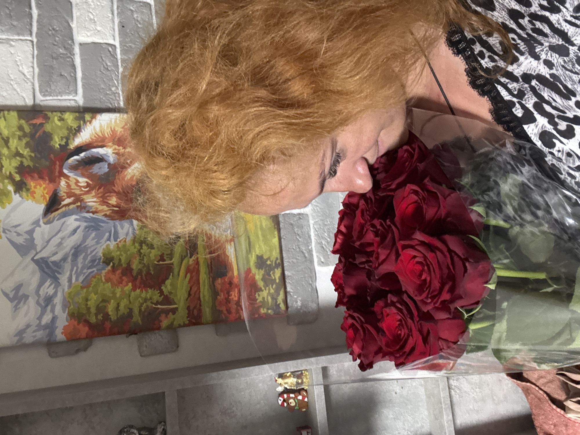 An indoor medium shot captures a woman with long, auburn hair holding a bouquet of red roses close to her face, appearing to smell them. She is looking down and to the left, with her eyes closed. The woman is wearing a black and white leopard-print top with black lace trim. In the background, to the left, a shelf unit holds small decorative items. Behind the woman, a framed picture depicts a landscape with mountains, trees with autumn foliage, and a fox. The wall behind the picture is made of grey and white bricks. The lighting suggests an indoor, domestic setting. The image was taken in the city of Chaikovsky, Russia.