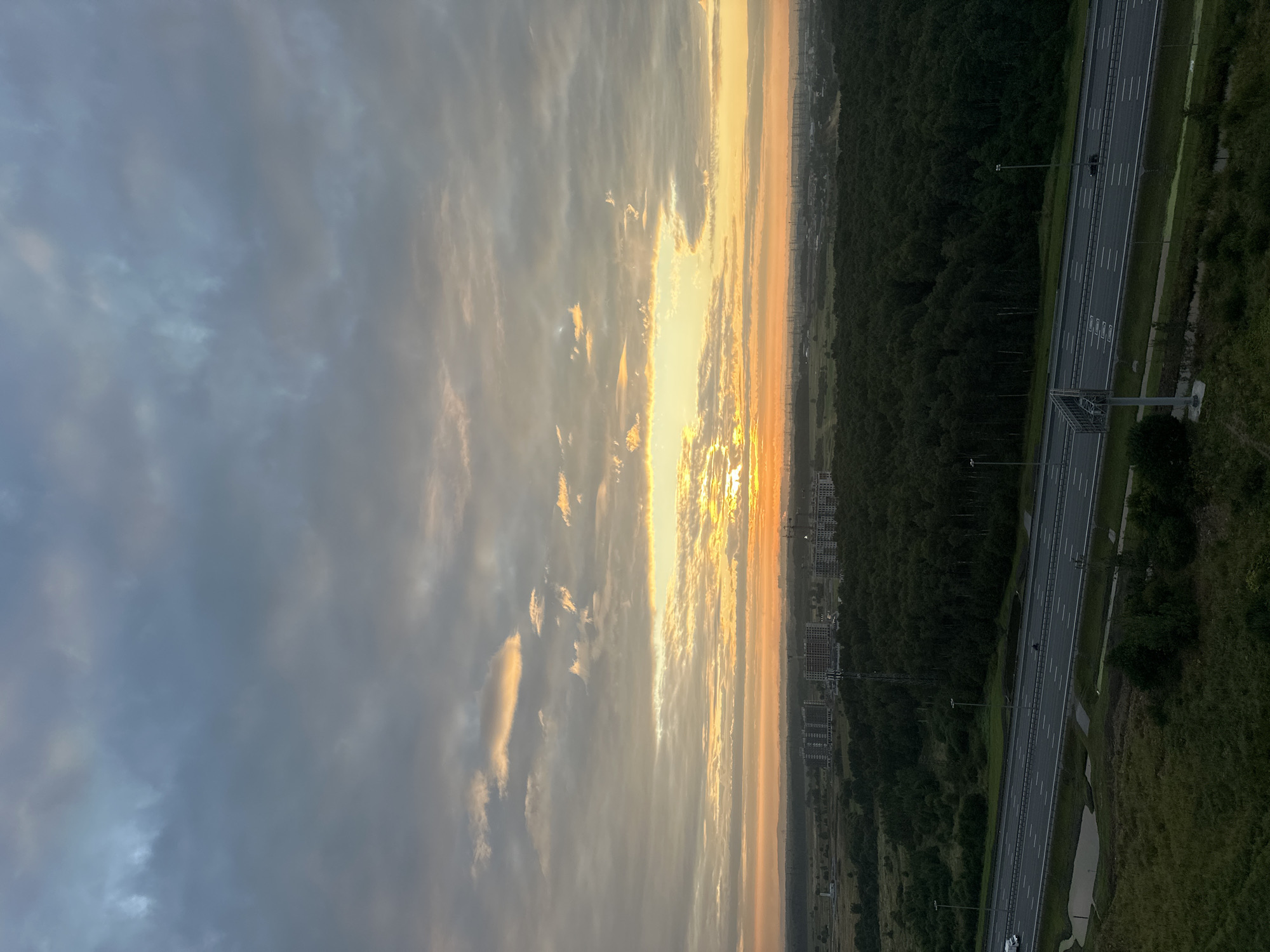 The image shows a wide-angle view of a suburban landscape at sunrise, likely in Kudrovo, Russia, given the characteristic construction sites and infrastructure. The sky is filled with a dramatic mix of gray, blue, and vibrant orange clouds, with the sun just breaking through on the horizon, casting a warm glow. In the middle ground, a dense, dark green forest stretches across the landscape, interrupted by several large, multi-story residential buildings, some appearing to be under construction with visible cranes. A multi-lane highway runs through the foreground, with a few vehicles, including a white van and a dark-colored car, visible on the road. The highway is bordered by grassy areas and streetlights. The overall scene conveys a sense of dawn breaking over a developing urban fringe, with a contrast between natural woodland and ongoing construction.