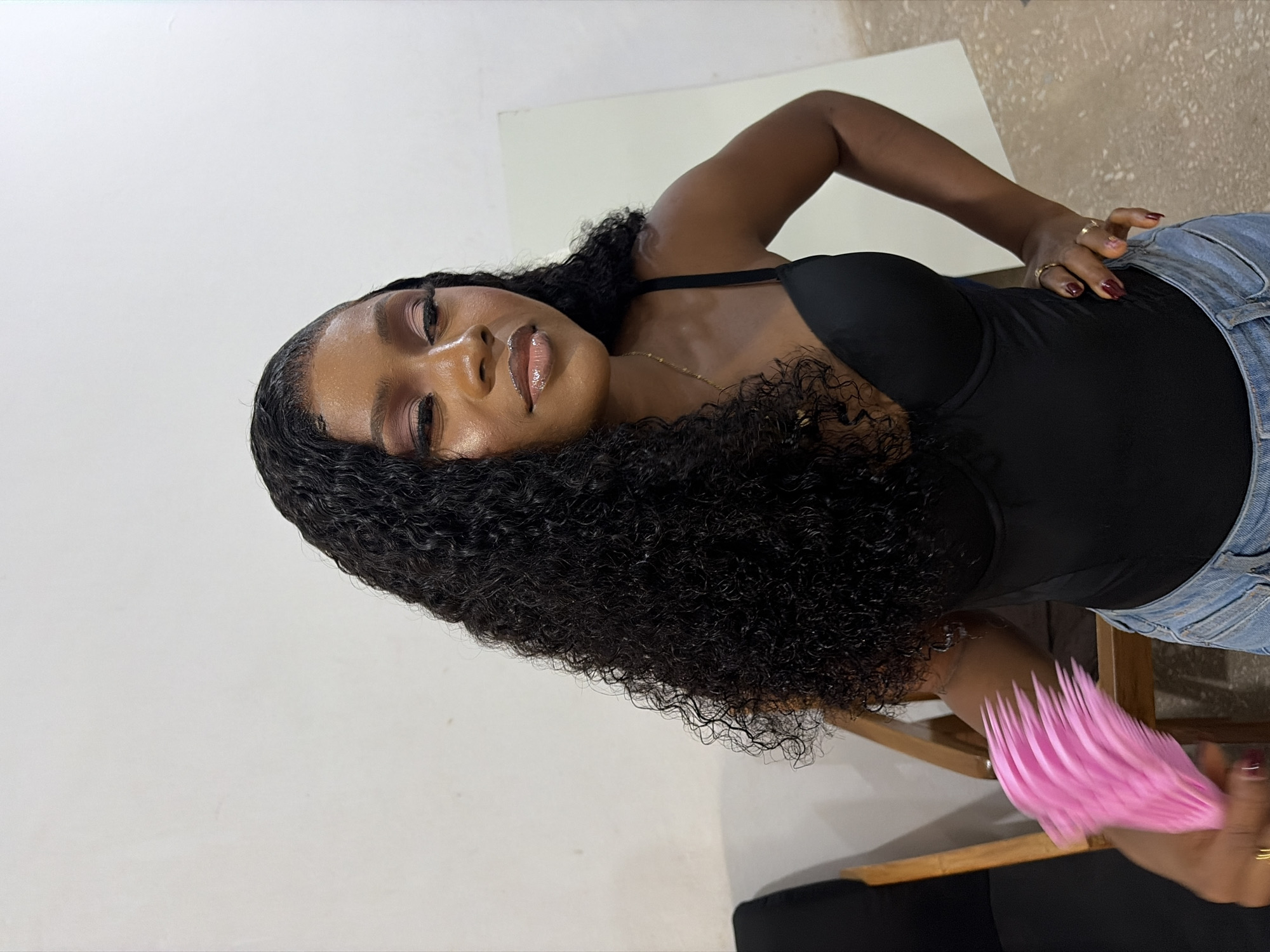 A young adult woman with dark skin and long, dark, curly hair stands indoors, posing against a plain light-colored wall in Gura Top, Nigeria. She wears a black sleeveless top with thin straps and light-wash denim jeans. Her face is made up with pink eyeshadow, winged eyeliner, false eyelashes, and glossy lips. A thin gold chain necklace with a small pendant and multiple gold rings are visible on her fingers. In her left hand, which is slightly blurred from motion, she holds a cluster of bright pink, thin, flexible objects, possibly a small fan or decorative item. Her right hand is placed on her hip. To her left, a dark chair with a wooden frame is partially visible, and to her right, a white panel or board leans against the wall. The floor is a light-colored concrete or tile.