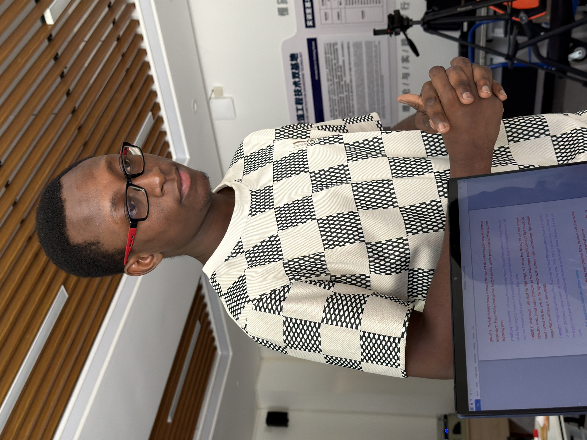 In a well-lit indoor environment, a young Black man wearing glasses with red accents and a checkered shirt stands partially turned to his right, facing the camera at a three-quarter angle. His hands are clasped in front of him. To his left, a laptop screen displays text. In the background, a poster with Chinese text is visible, along with part of a tripod and what appears to be part of a chair or stand. The ceiling features a modern design with wooden slats and recessed lighting. The setting suggests a presentation or discussion, potentially within an academic or research institution, given the poster and the presence of the laptop and tripod, which could be used for recording or presentation purposes. The overall impression is one of a professional or educational setting. The location is identified as Jiangjin, China.