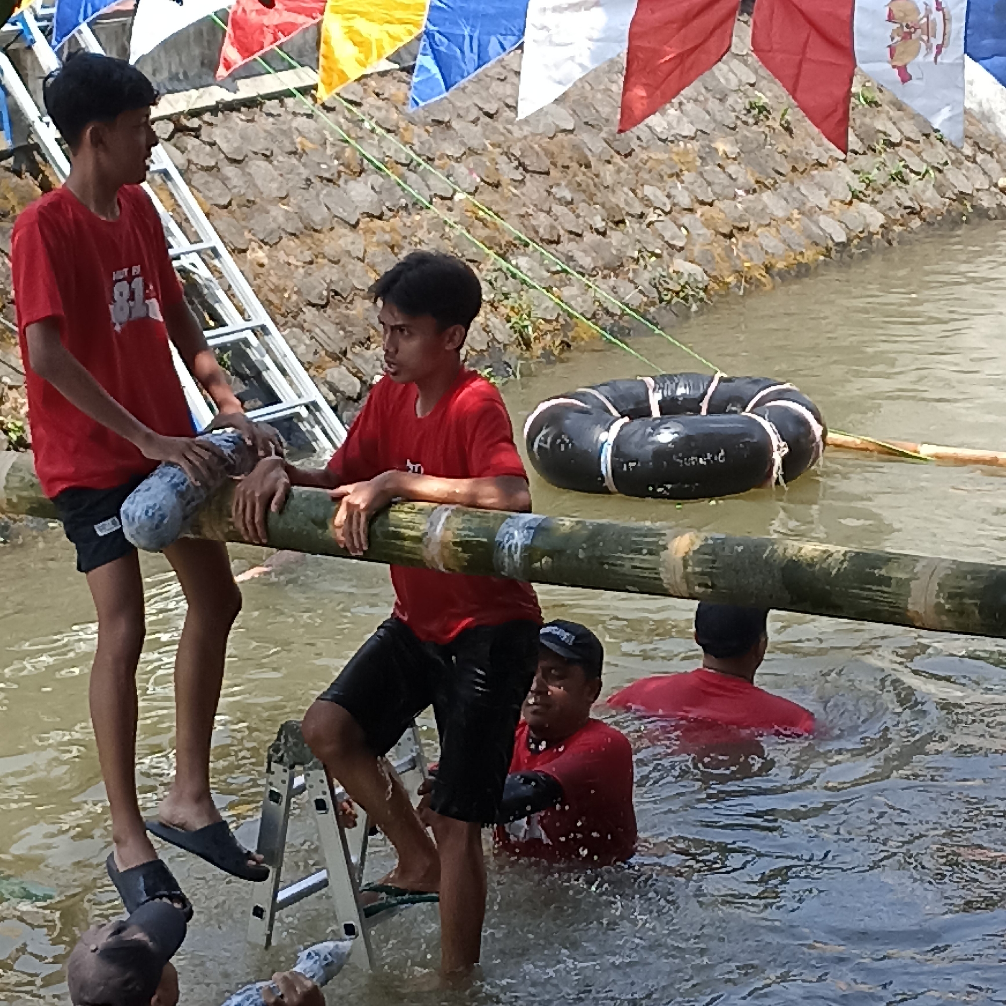 In the city of Magelang, Indonesia, a group of young men are participating in a water-based game, likely a traditional event given the festive flags adorning the background. The primary focus is on two individuals, one slightly taller than the other, both wearing red t-shirts and dark shorts. The taller youth is positioned on a bamboo pole, holding it with both hands. His companion, standing next to him on the same pole, is also holding it, with one hand on the pole and the other seemingly supporting a grey, cylindrical object wrapped around the bamboo. Both are immersed in murky water, with splashes indicating movement. A metal ladder is partially visible in the water, suggesting it is used for access to the bamboo pole or as part of the game. Further in the background, another young man in a red t-shirt is partially submerged, looking towards the action. Two black inflatable rings, connected by ropes, float near the bamboo pole. The environment appears to be a river or canal, with a stone-lined bank and lush green vegetation on the embankment. The overall atmosphere is one of active participation and community engagement.