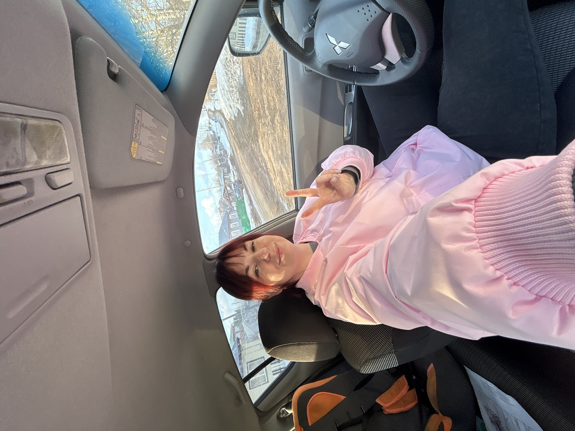 A woman in a pink jacket is sitting in the driver's seat of a Mitsubishi car, holding up two fingers in a "peace" sign. The view from the car window shows a snowy streetscape with houses, suggesting the location is in a residential area. The dashboard, steering wheel with the Mitsubishi logo, and sun visor are visible inside the car. A backpack is resting on the passenger seat. The image was taken in Фоки, Russia.