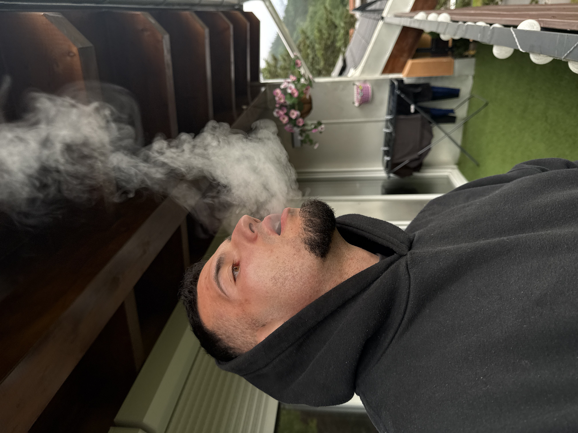 A man exhales a cloud of smoke from his mouth, his face angled upwards. He is wearing a black hooded sweatshirt. The man appears to be standing on a balcony with artificial turf flooring. In the background, a clothes drying rack with clothes, a hanging basket of pink flowers, and a railing with decorative lights are visible. The surrounding environment suggests an outdoor setting, possibly a residential area in Garmisch-Partenkirchen, Germany, with buildings and trees in the distance.