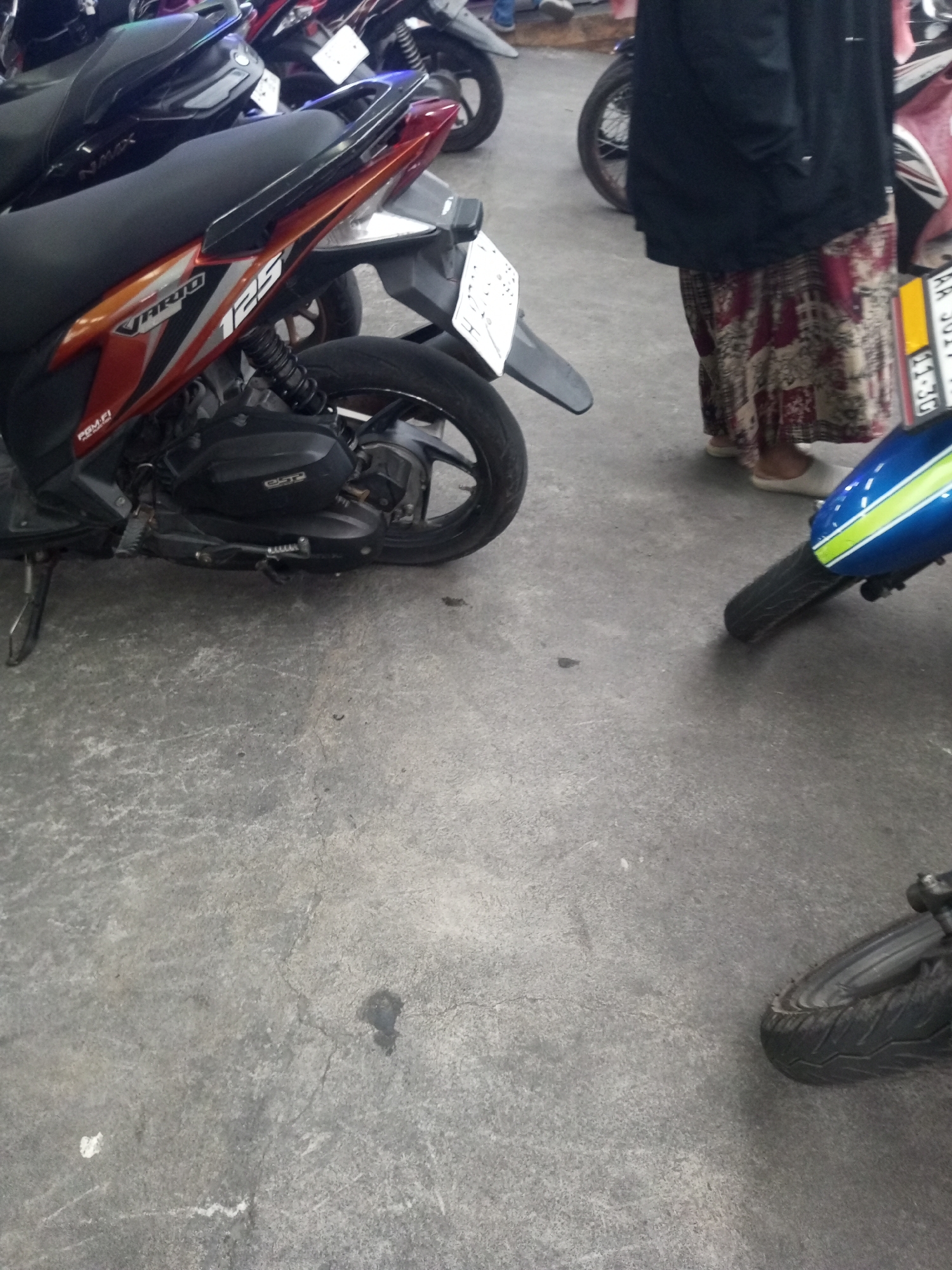 The image depicts a street scene in Kalibeber, Indonesia, with a focus on parked motorcycles and a person. A red and black scooter is prominently featured in the foreground, with its rear tire appearing flat. To the right, a person wearing a black jacket and a patterned skirt stands near the front wheel of a blue and green motorcycle. The ground is concrete, showing some wear and tear with a few dark smudges and lighter markings. Other motorcycles are visible in the background, also parked on the concrete surface. The lighting suggests it is daytime, with shadows cast on the ground.