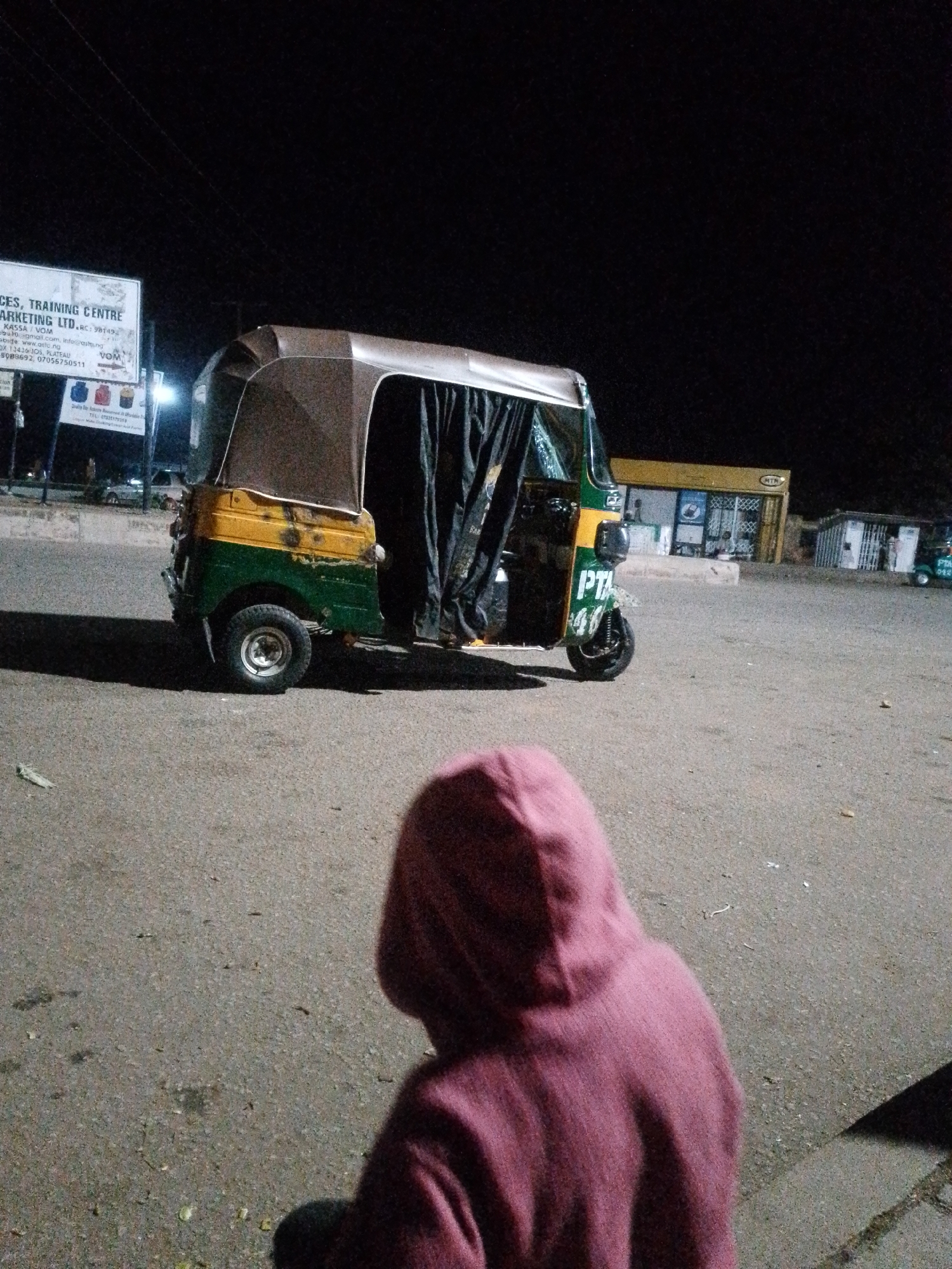 The image captures a night scene in Bukuru, Nigeria, with a yellow and green auto-rickshaw parked on an asphalt surface. The auto-rickshaw is partially covered with dark curtains, and the word "PTA" is visible on its side. In the foreground, a person wearing a reddish-purple hooded sweatshirt is partially visible, with their back to the camera.
In the background, several buildings are illuminated. One building appears to be a shop or store with yellow siding and display windows. Another structure to the right looks like a smaller building with barred windows. A sign is visible on the left side of the image, partially obscured by darkness, with text that includes "CES, TRAINING CENTRE" and "MARKETING LTD". The sky is dark, indicating nighttime. The ground is a rough asphalt surface, with some scattered debris.