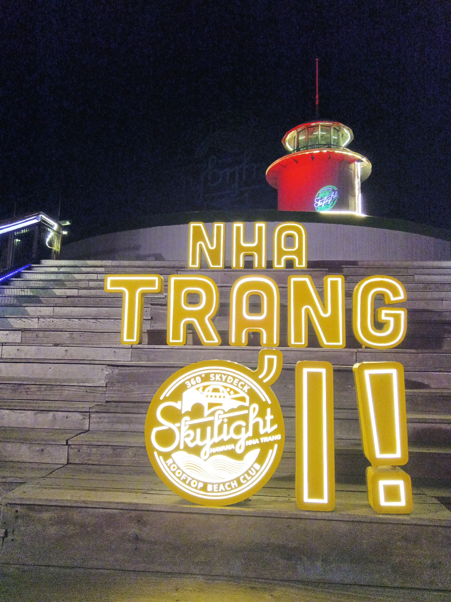 The image shows a lit sign spelling "NHA TRANG" in large yellow block letters on a staircase. Below it is a circular sign for "Skylight 360° Skydeck Havana Nha Trang Rooftop Beach Club," also illuminated in yellow. To the right of the circle are two large yellow exclamation points. Behind the signs is a red and white building structure with a red pole on top and a "Skylight" logo. The scene appears to be at night in Nha Trang Ward, Vietnam.