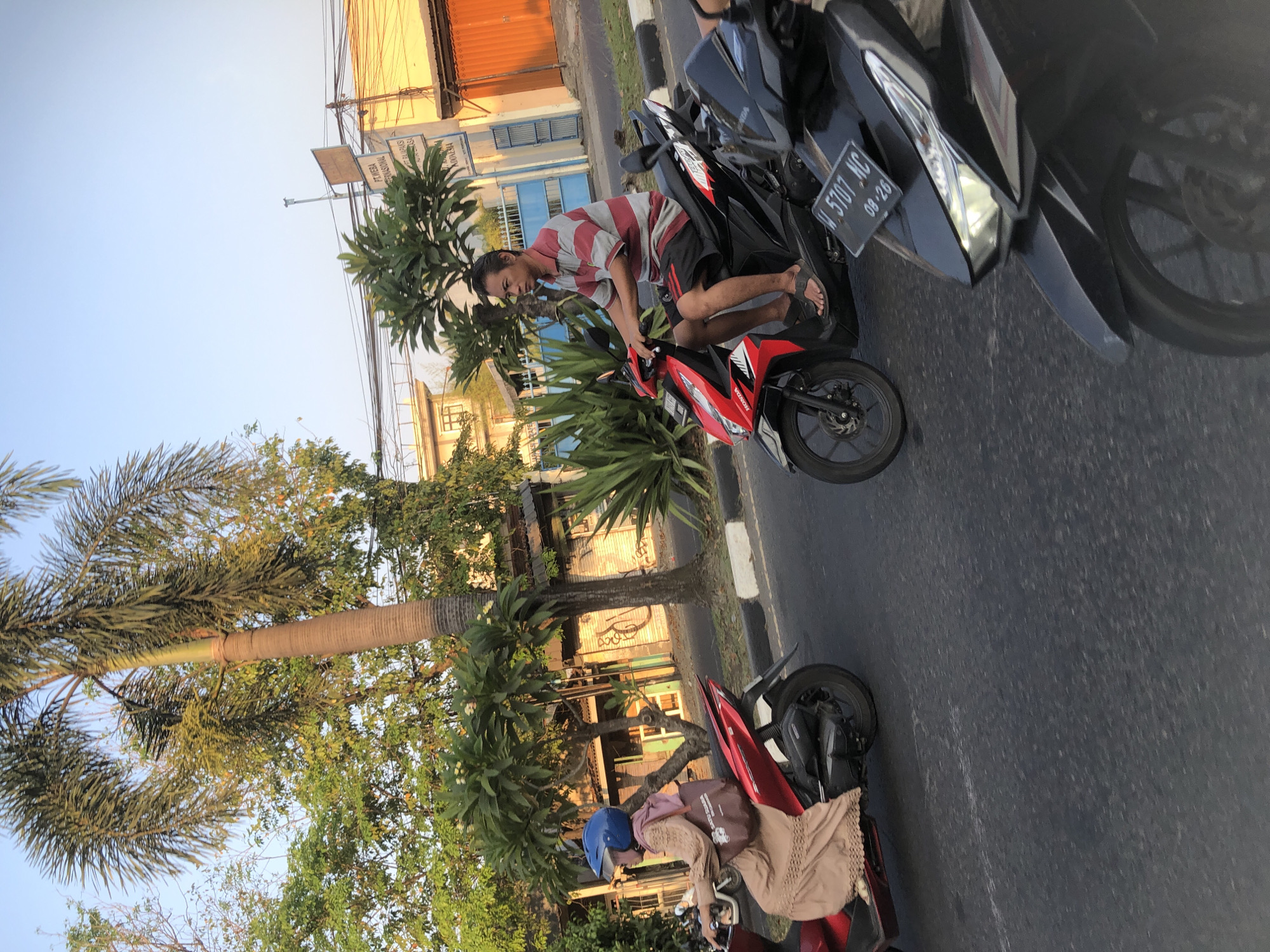 This image captures a street scene in Candi, Indonesia, likely during the late afternoon or early evening, given the warm lighting. Two motorcycles are in motion on a paved road. The motorcycle on the left is red and is ridden by a woman wearing a light brown abaya and a blue helmet. She is carrying a dark brown tote bag on her shoulder. The motorcycle on the right is a dark color, possibly black, and is ridden by a man wearing a striped t-shirt and black shorts. He is wearing sandals. The foreground shows the front of the black motorcycle, with a license plate reading "W. 5707 NC 08-26".
The environment includes a sidewalk with white markings and greenery. In the background, there are buildings with yellowish and orange facades, some with blue gates and fences. Lush trees, including a tall palm tree, are present, with dense foliage creating a backdrop. Power lines and streetlights are also visible, indicating an urban setting. The sky is a clear, light blue. The image appears to be taken from a moving vehicle, suggesting a candid street photography perspective.