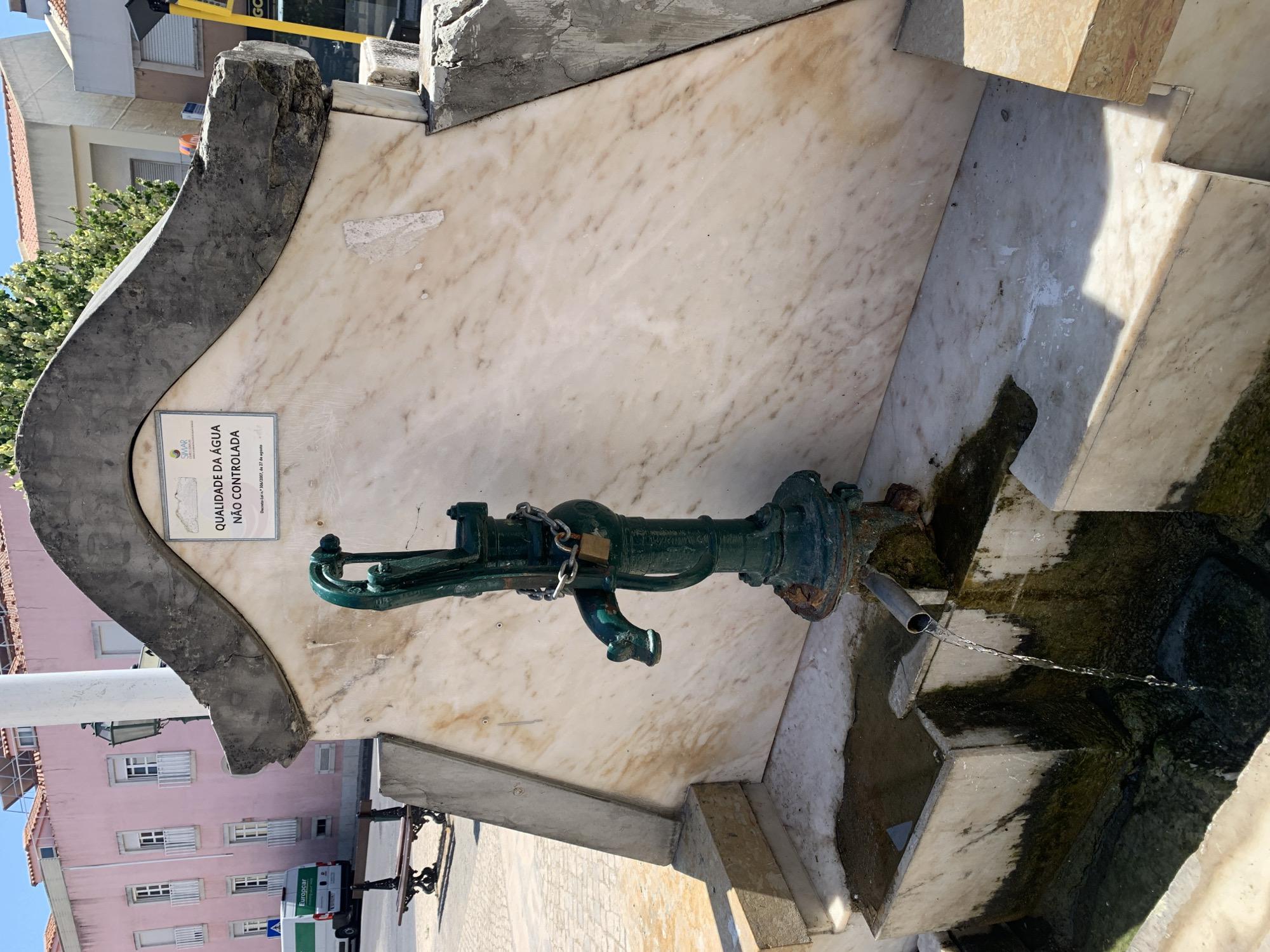 This is a photograph of an outdoor water pump in Odivelas, Portugal. The pump is green and made of metal, with a padlock and chain securing its handle. It is mounted on a marble structure that appears to be part of a fountain. Water is flowing from a spout into a basin below. The basin and surrounding marble are stained and covered in moss, suggesting the fountain is old or not well-maintained.
A sign on the marble structure reads "QUALIDADE DA ÁGUA NÃO CONTROLADA" which translates to "WATER QUALITY NOT CONTROLLED." Below this, it says "Decreto-Lei 306/2007, de 27 de agosto," referencing a Portuguese law regarding water quality.
In the background, a pink building with white-trimmed windows and balconies is visible. To the left of the pink building, there are trees. Further in the background, a yellow construction crane and a white van with "Europcar" branding are present.
The scene appears to be set during the daytime under clear skies, as indicated by the strong shadows. The overall impression is of a public space with a functional but weathered water source. The presence of the Europcar van suggests it is an urban or semi-urban environment.