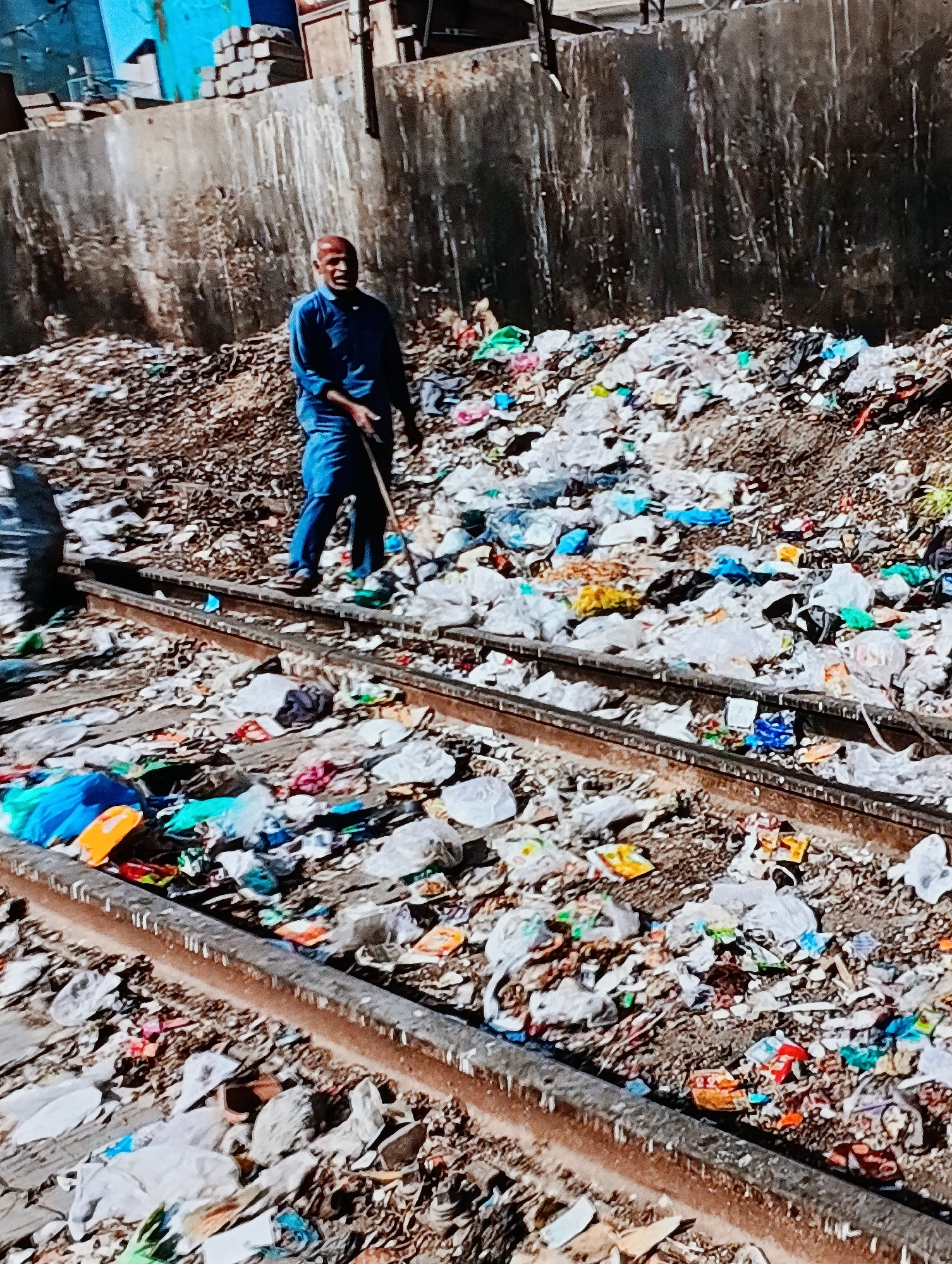 A man in a blue uniform and red cap stands on railroad tracks covered in garbage in an urban area. He holds a stick and is looking towards the viewer. The tracks are embedded in a large pile of garbage, mostly plastic bags and other debris. In the background is a high concrete wall, and beyond that, some buildings. The location appears to be in the city of Unknown, Unknown.