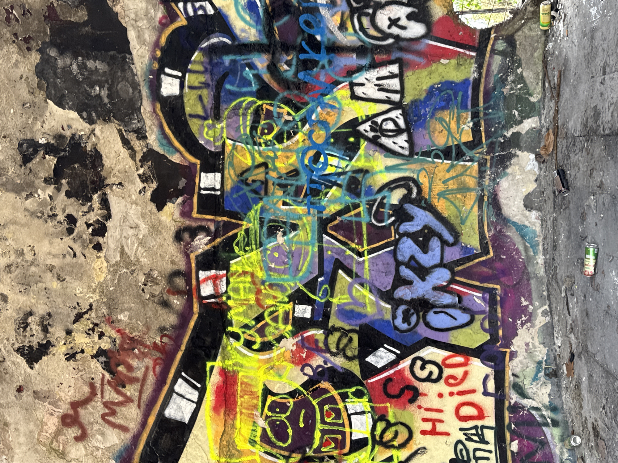 The image shows graffiti on a concrete wall. The wall is damaged and shows signs of deterioration with chipped paint and exposed concrete. The graffiti is layered and colorful, with a variety of shapes, lines, and letters in yellow, blue, black, white, purple, and red. The graffiti appears to be abstract and contains some discernible words, including "Hi Died". Two empty spray paint cans are resting on the concrete, right of the graffiti. The exact location in Varna, Bulgaria is not indicated by the image. There are no people visible.