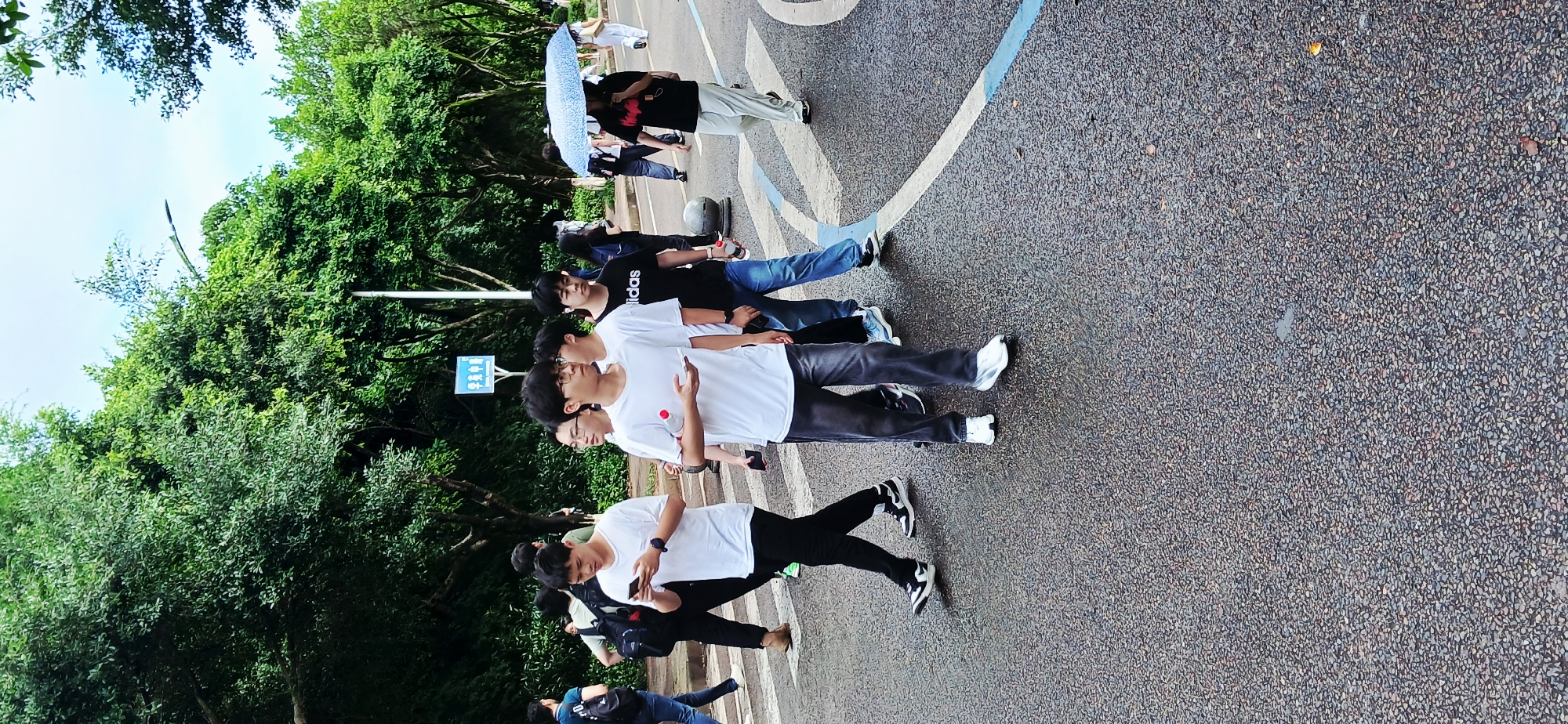 A group of people is standing on a road with a white and blue painted design in Chongqing High-tech Zone, China. The road surface is textured, possibly asphalt or concrete. Trees line the edge of the road. The people are wearing a mix of casual clothing, including t-shirts, jeans, and sneakers. Some are using mobile phones. One person is holding an umbrella. A blue sign with white lettering is visible near the trees.