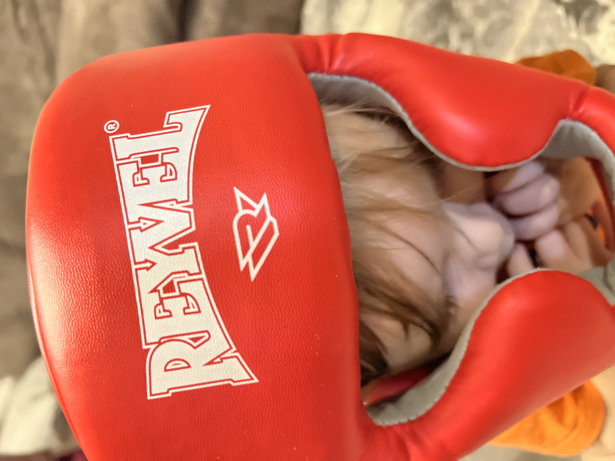 A red boxing headgear with the word "REVEL" in white is prominently displayed. The headgear is partially obscuring a person's face, with some hair visible. The background is indistinct. The location is not discernible from the image.