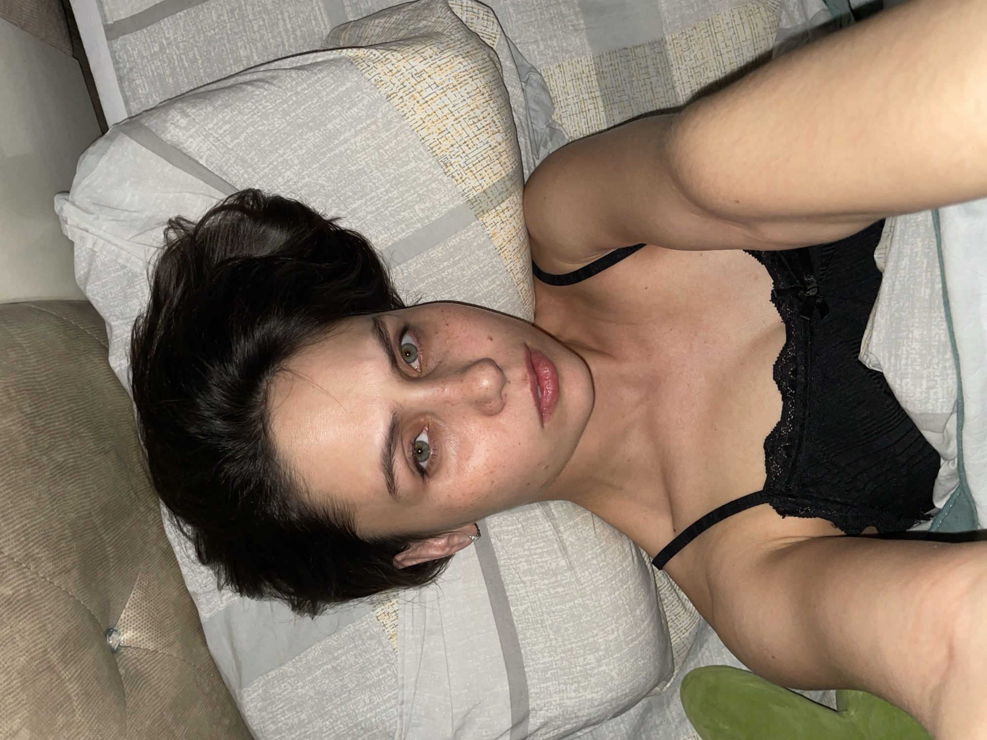 A woman lies on a pillow with a patterned cover. She has dark hair pulled back, fair skin, and visible freckles on her face. She wears a black lace top with thin straps. Her head is turned slightly toward the camera. The setting appears to be a bed, with a couch visible in the background. There is no information to indicate that this image was taken in Izhevsk, Russia.