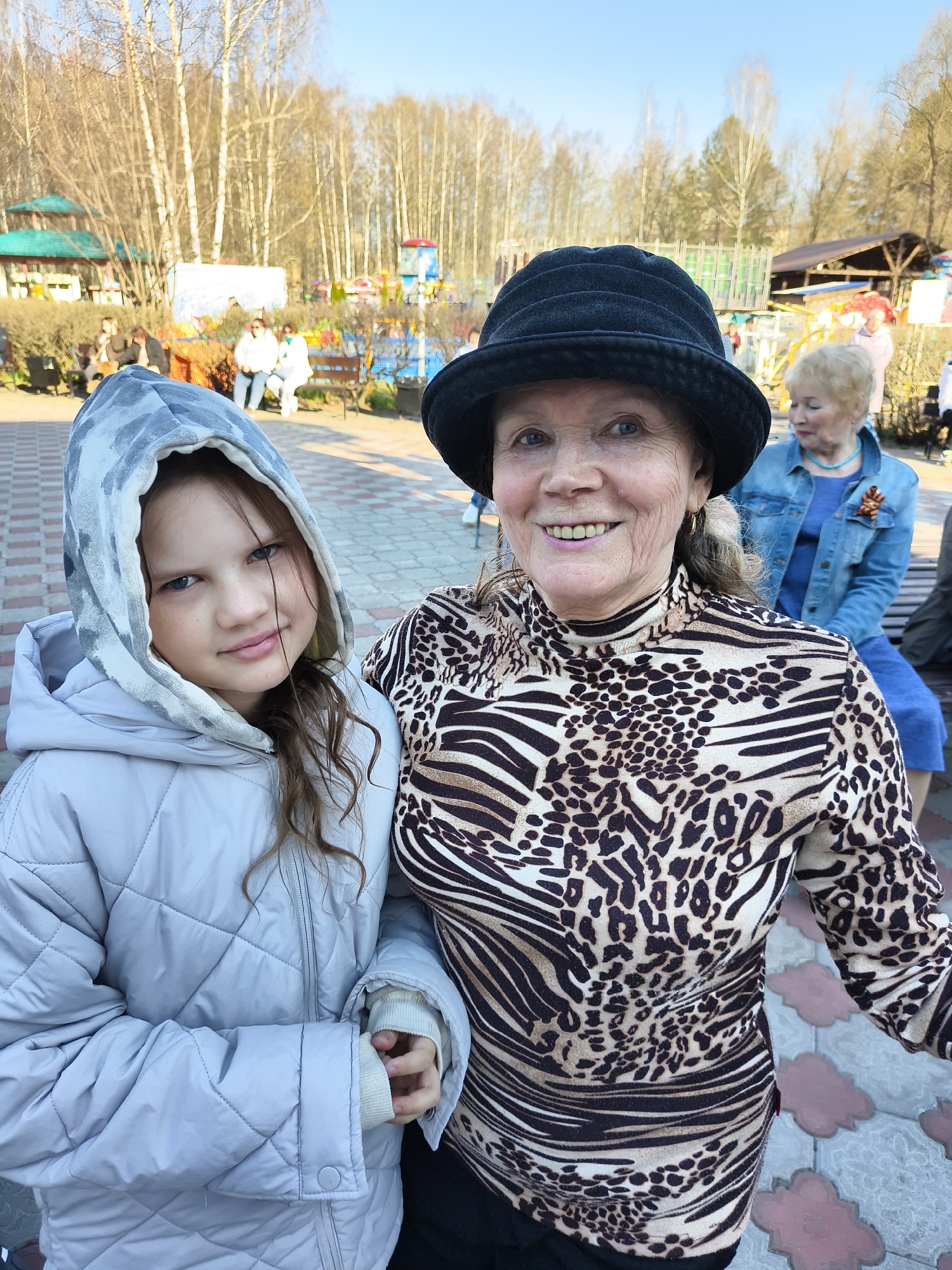 The image features a young girl and an older woman, possibly grandmother and granddaughter, posing for a photo in an outdoor setting in a city. The girl, on the left, wears a light grey quilted jacket with a hood up. Her hair is dark brown. The older woman, on the right, wears a black hat and a long-sleeved animal print top. Both are looking at the camera and smiling.
In the background, there is a brick-paved area with benches. Other people can be seen in the distance. There are also trees and some colorful structures that could be part of a park or a playground.